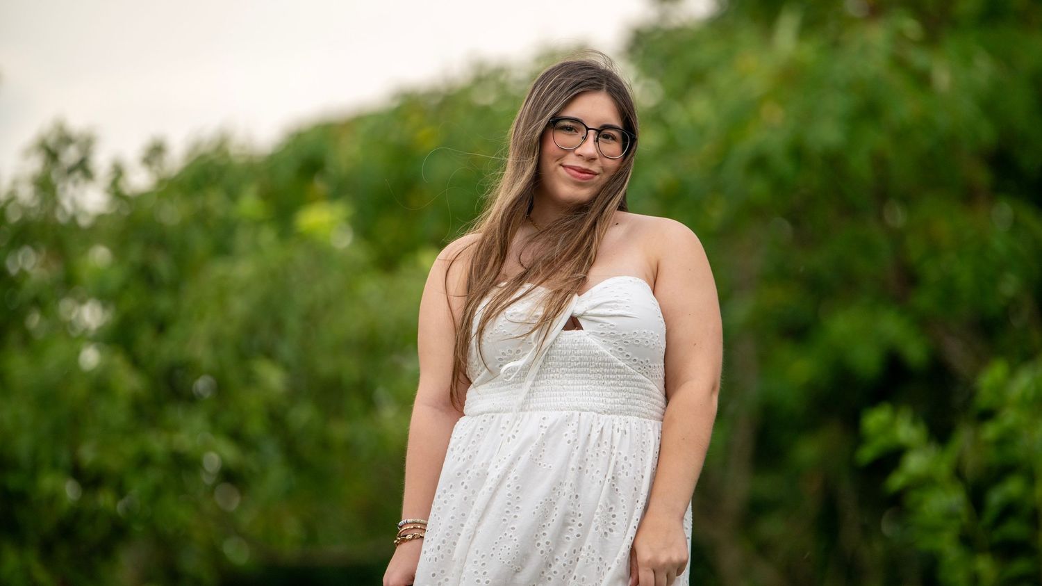 A person in a strapless white dress stands in front of green trees.