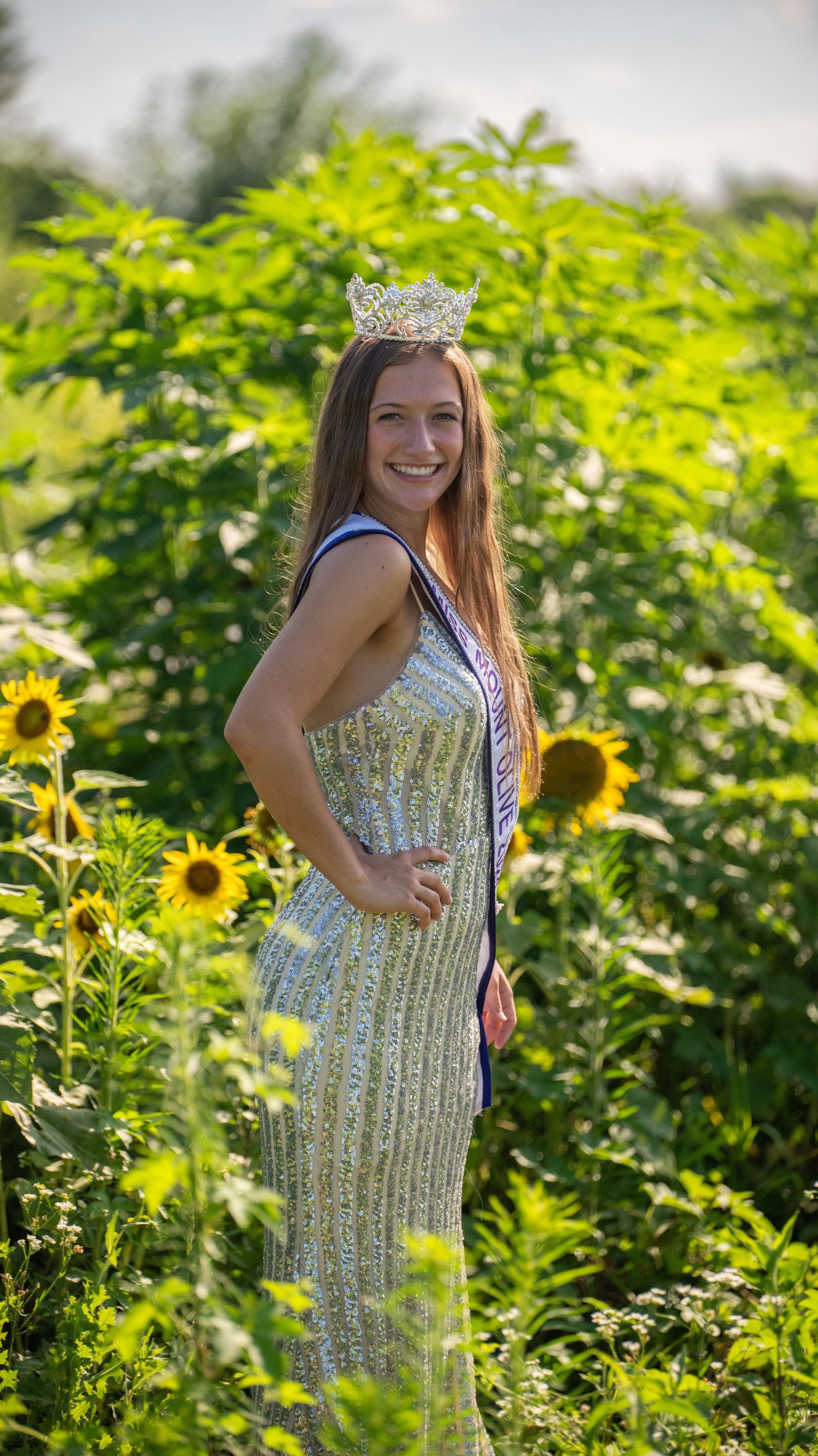 A crowned pageant contestant poses in a sequined dress among blooming sunflowers.