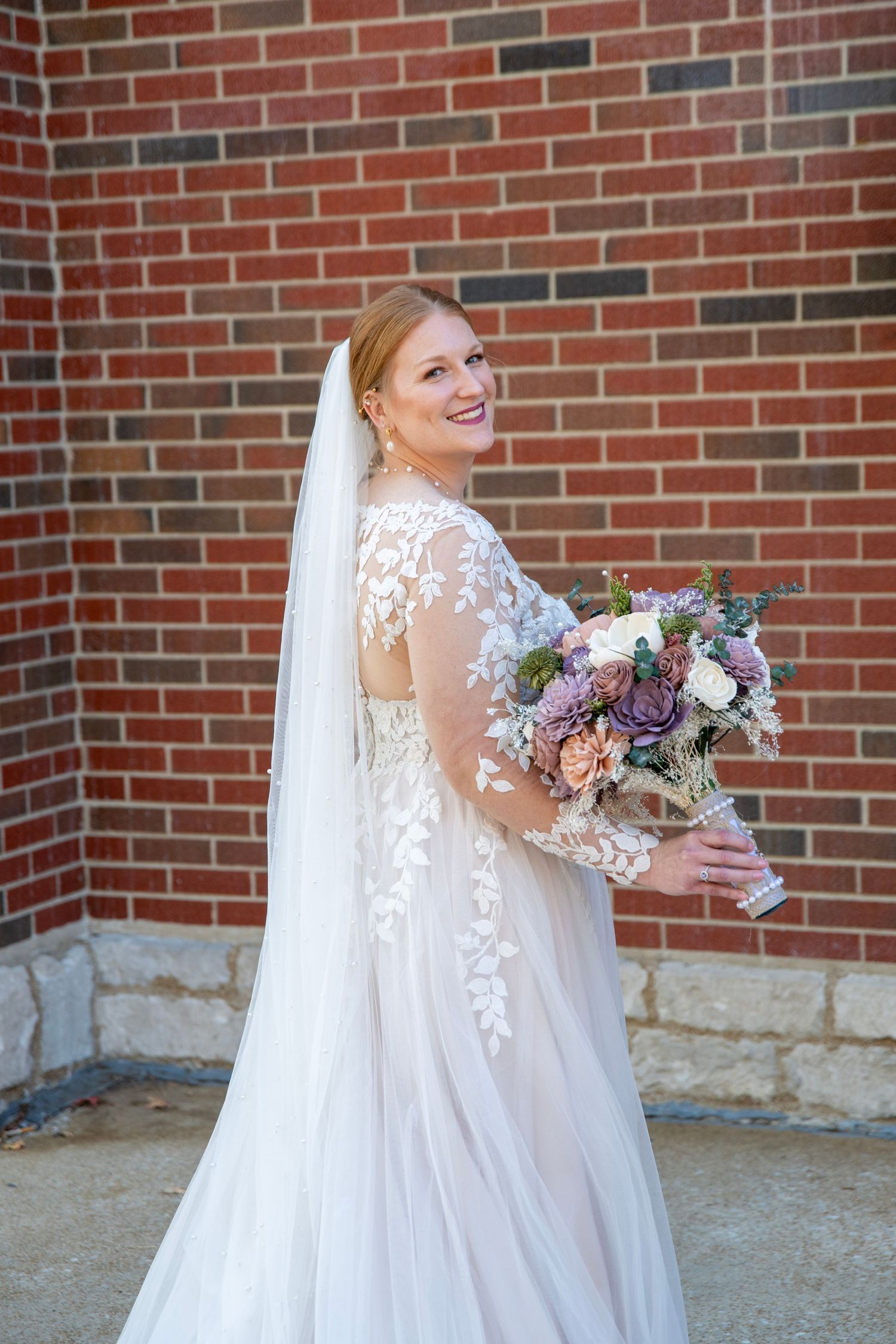 Bride in elegant lace gown holds a purple and pink bouquet against brick church backdrop.