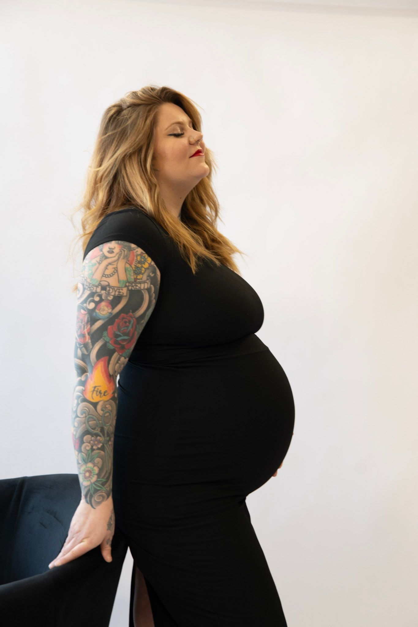 Woman in black dress showing off colorful sleeve tattoos in maternity photoshoot.
