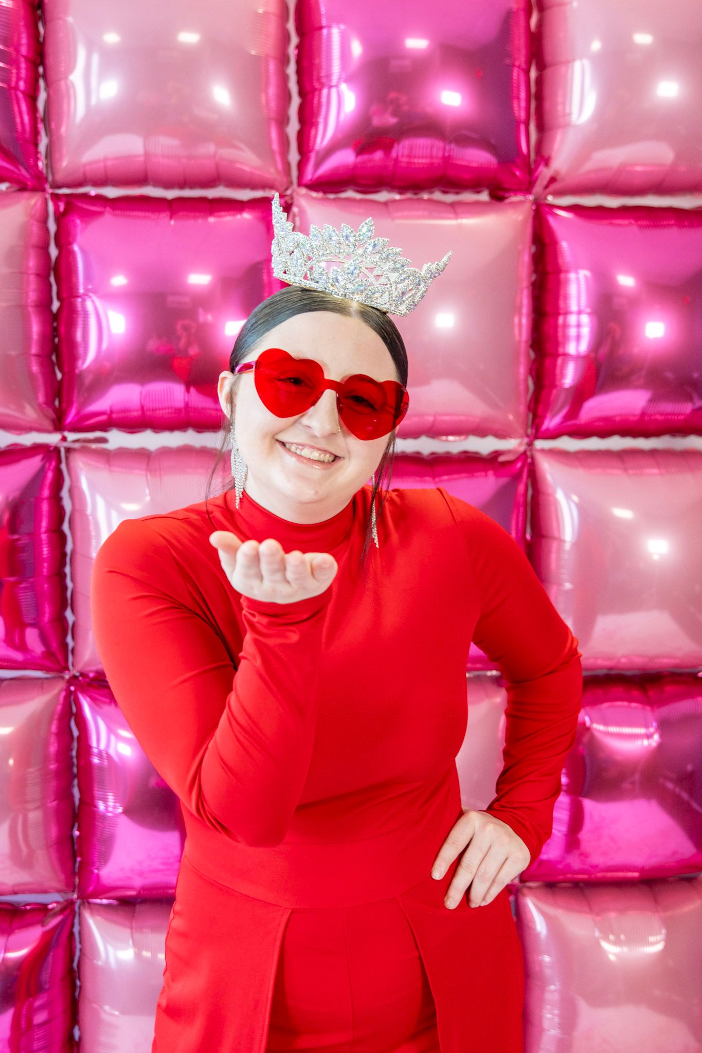 Red outfit and heart sunglasses against pink metallic balloon wall backdrop.