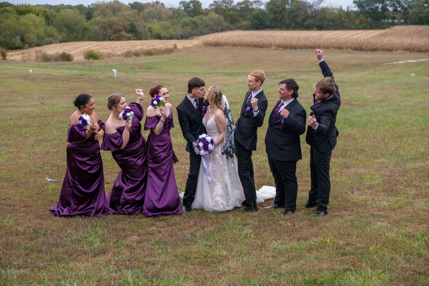 Wedding party gathered in a line on grassy hillside wearing purple dresses and dark suits.