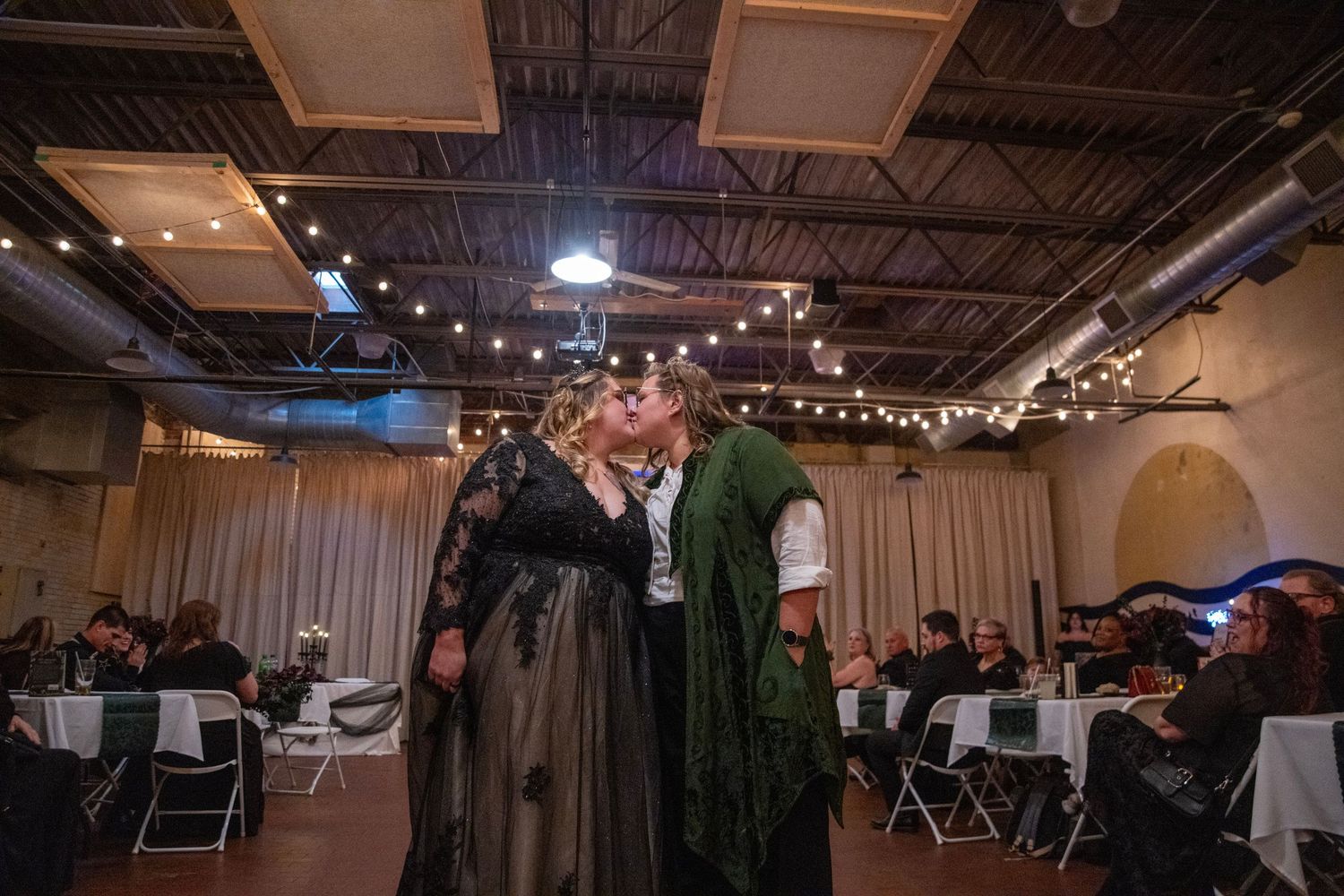 Two people in dark Victorian-style costumes embracing at an indoor event venue.