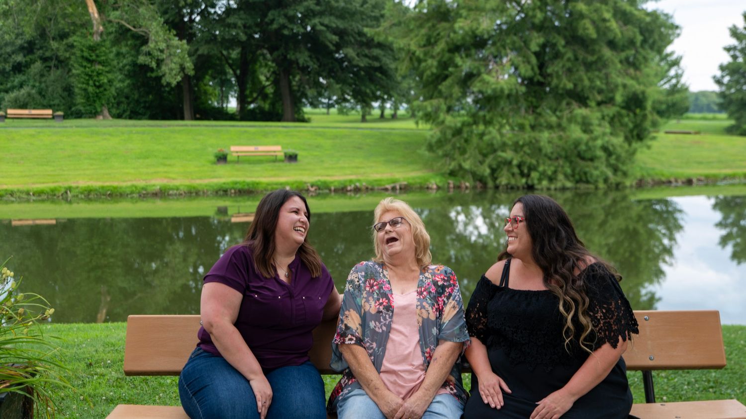 Three friends share joyful laughter while sitting together on a bench by a serene pond.