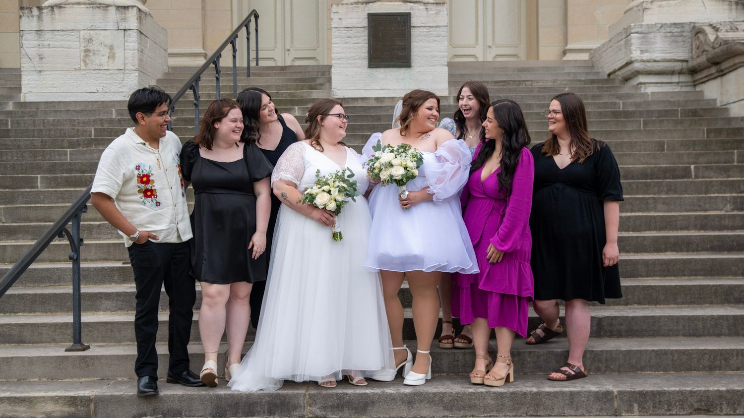 A wedding party group poses on stone steps wearing mix of formal attire in black and bright colors.