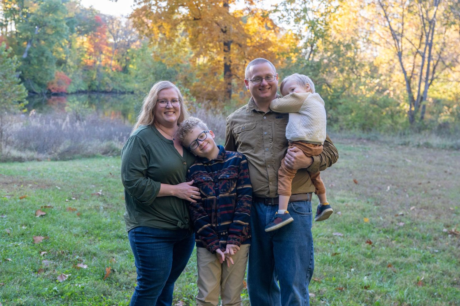 Family members embrace while standing in a grassy field surrounded by autumn trees.