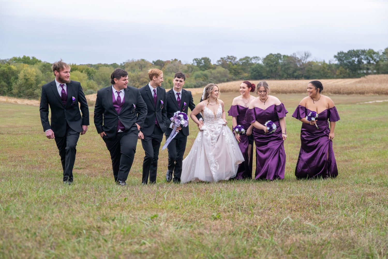Wedding party walking together across grassy field in coordinated purple and black attire.