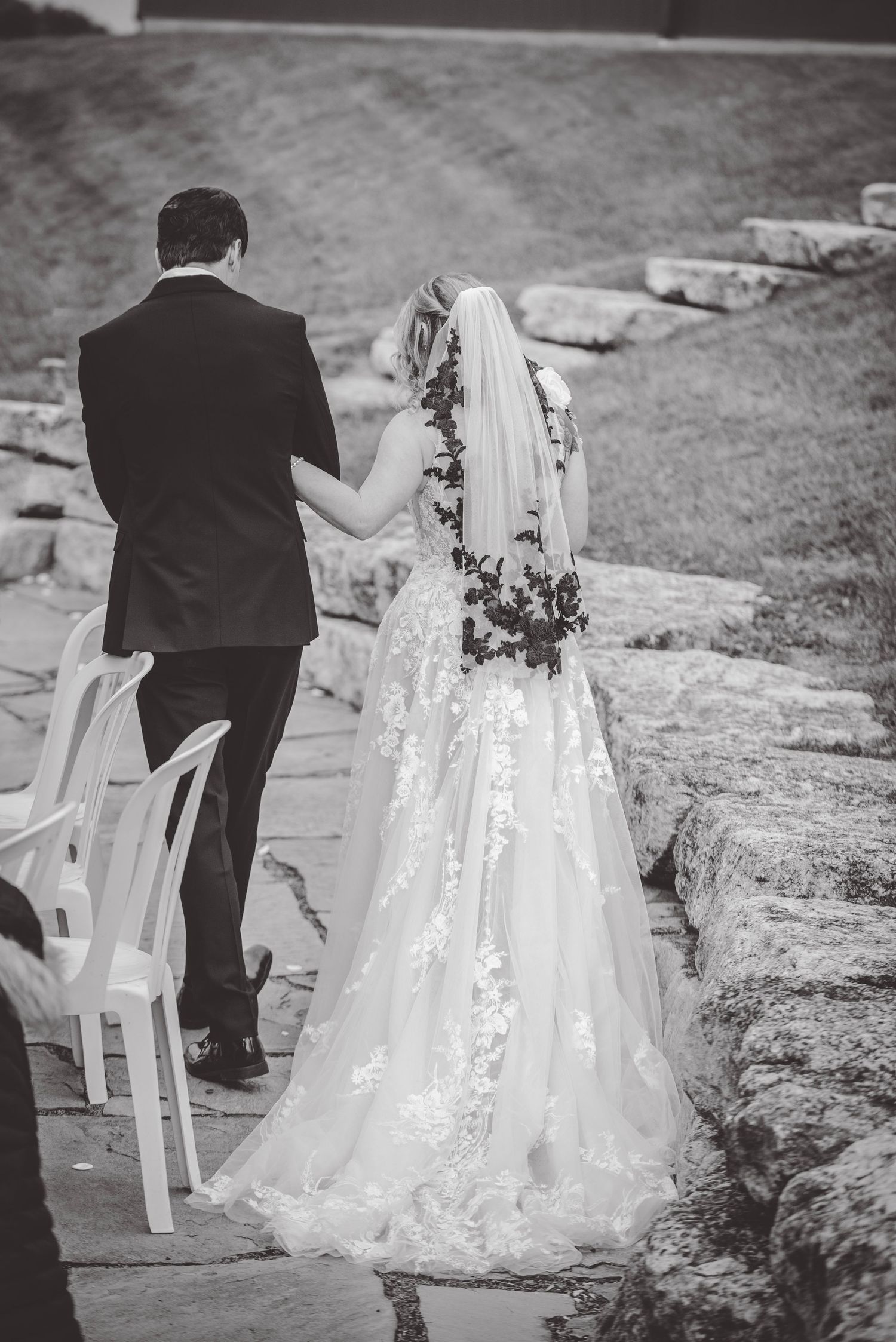 Black and white photo of wedding couple walking along stone pathway with white chairs.