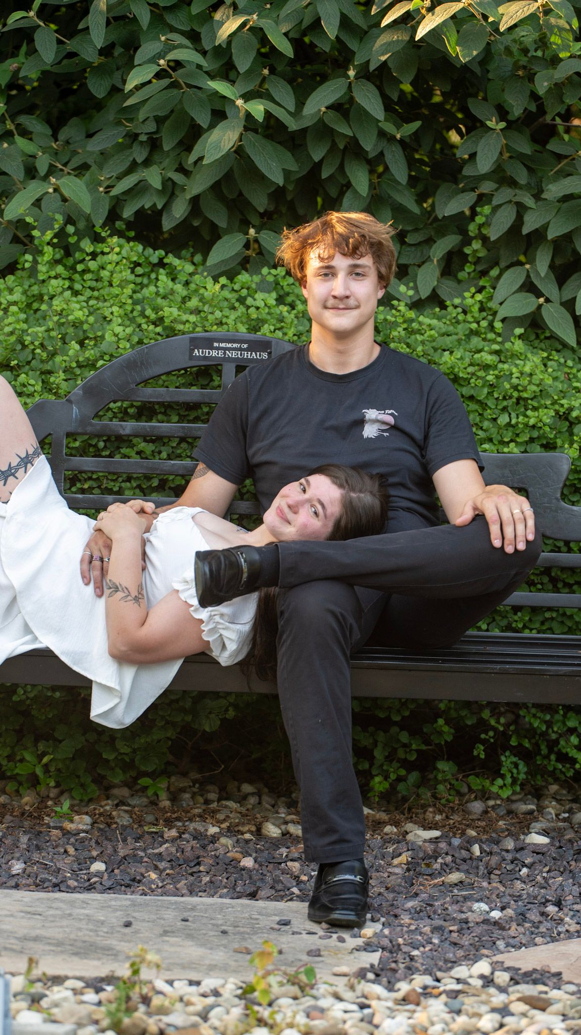 A couple shares a relaxed moment on a black garden bench surrounded by greenery.