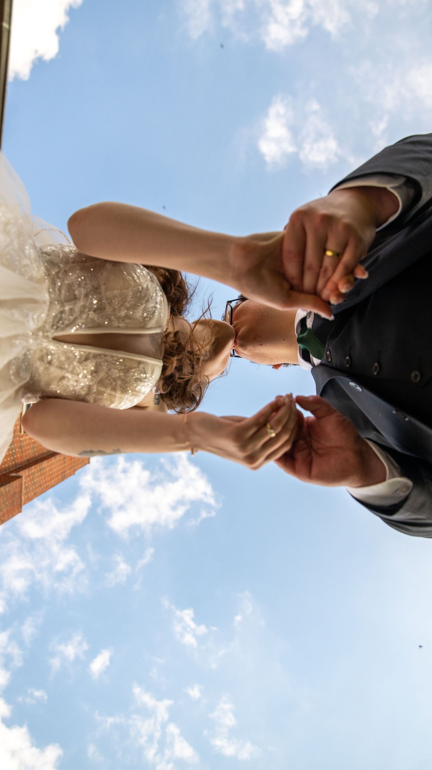 A creative upward angle shot of a couple holding hands against a blue sky with clouds.