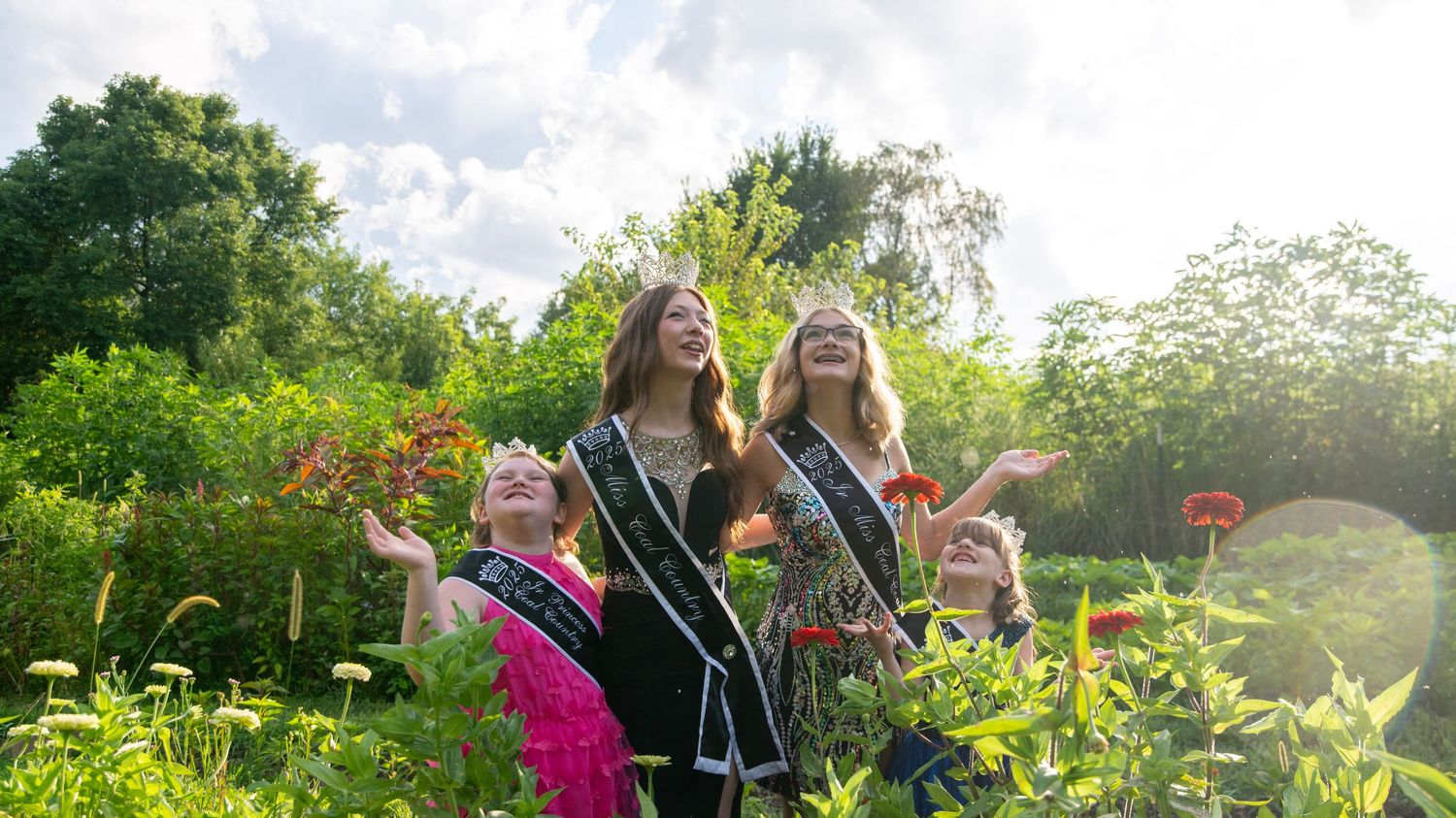 Group wearing sashes and crowns pose joyfully among blooming garden flowers.