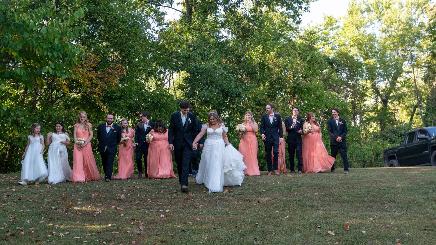 Wedding party members stand in a line outdoors under green trees on a sunny day.