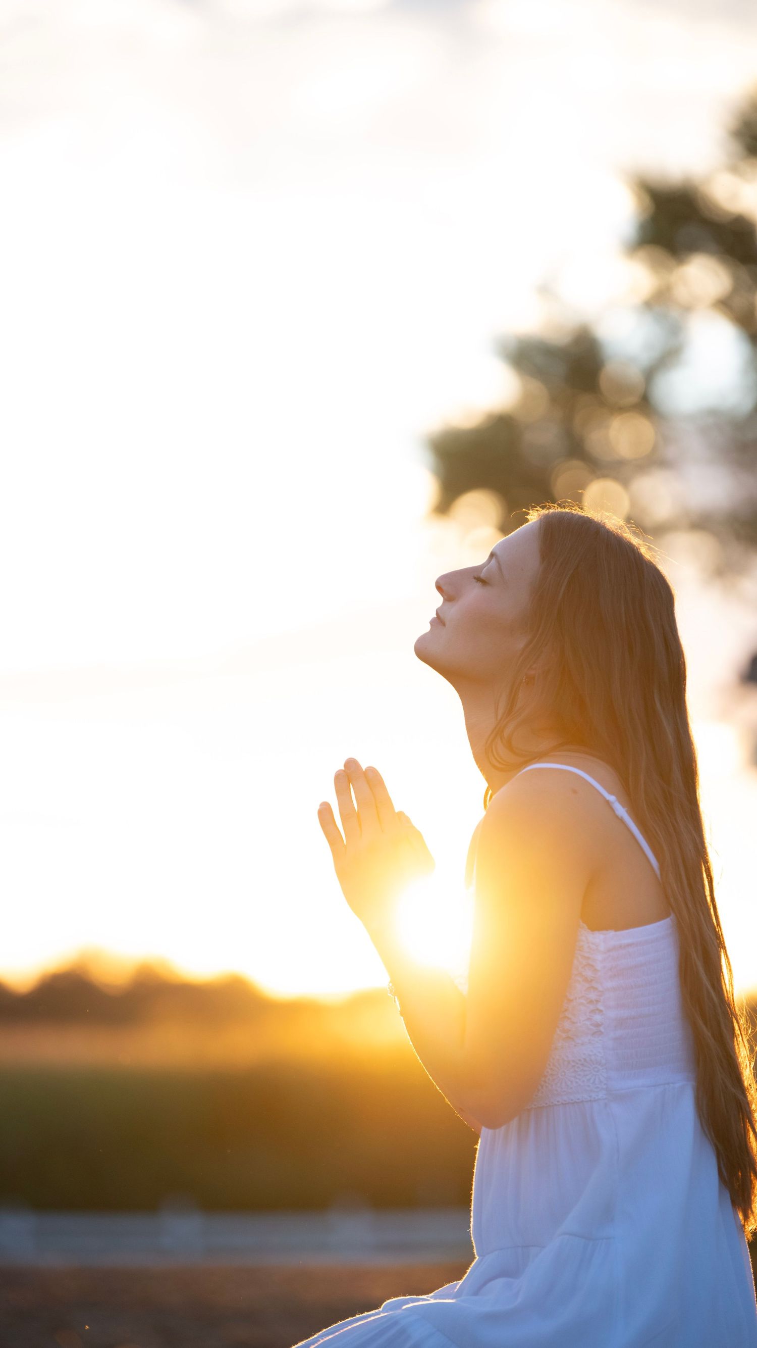 Silhouetted figure in white dress praying during golden hour sunset.