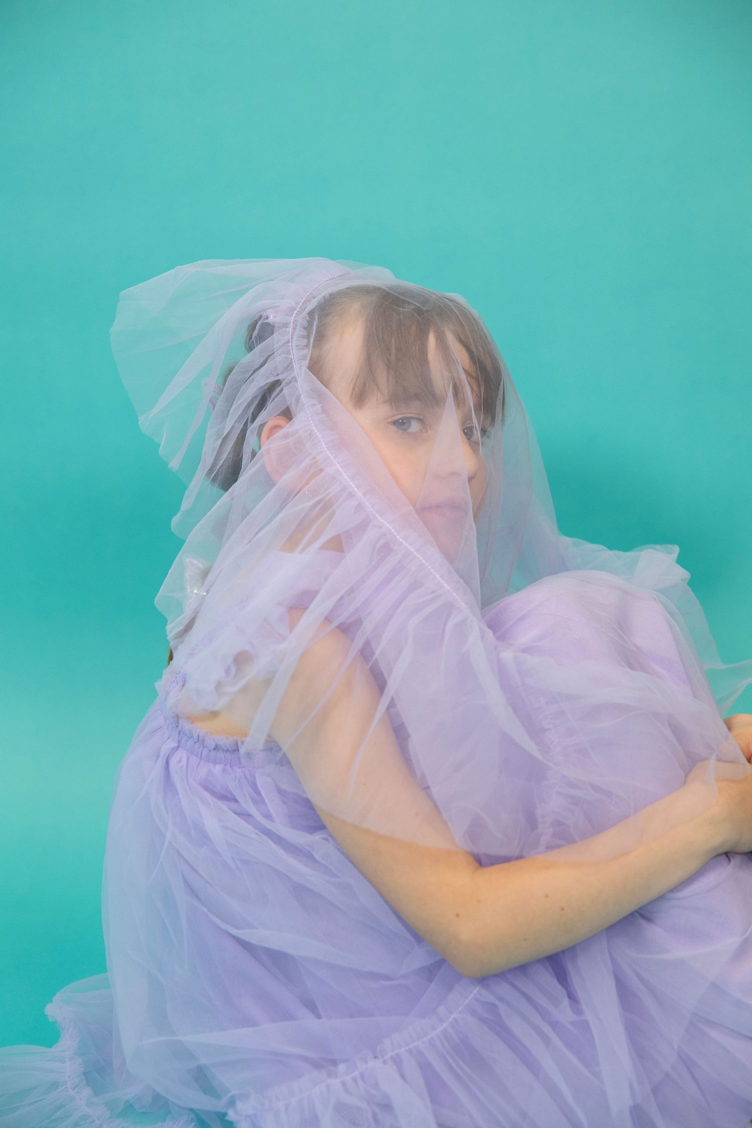 A figure in a lavender tulle dress poses playfully against a turquoise backdrop.