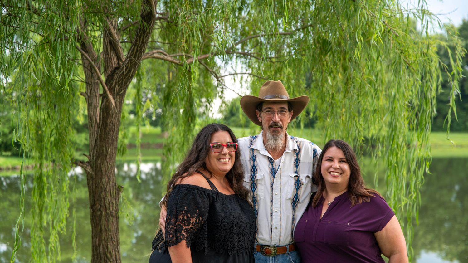 Three people stand together under a weeping willow tree near a peaceful pond.