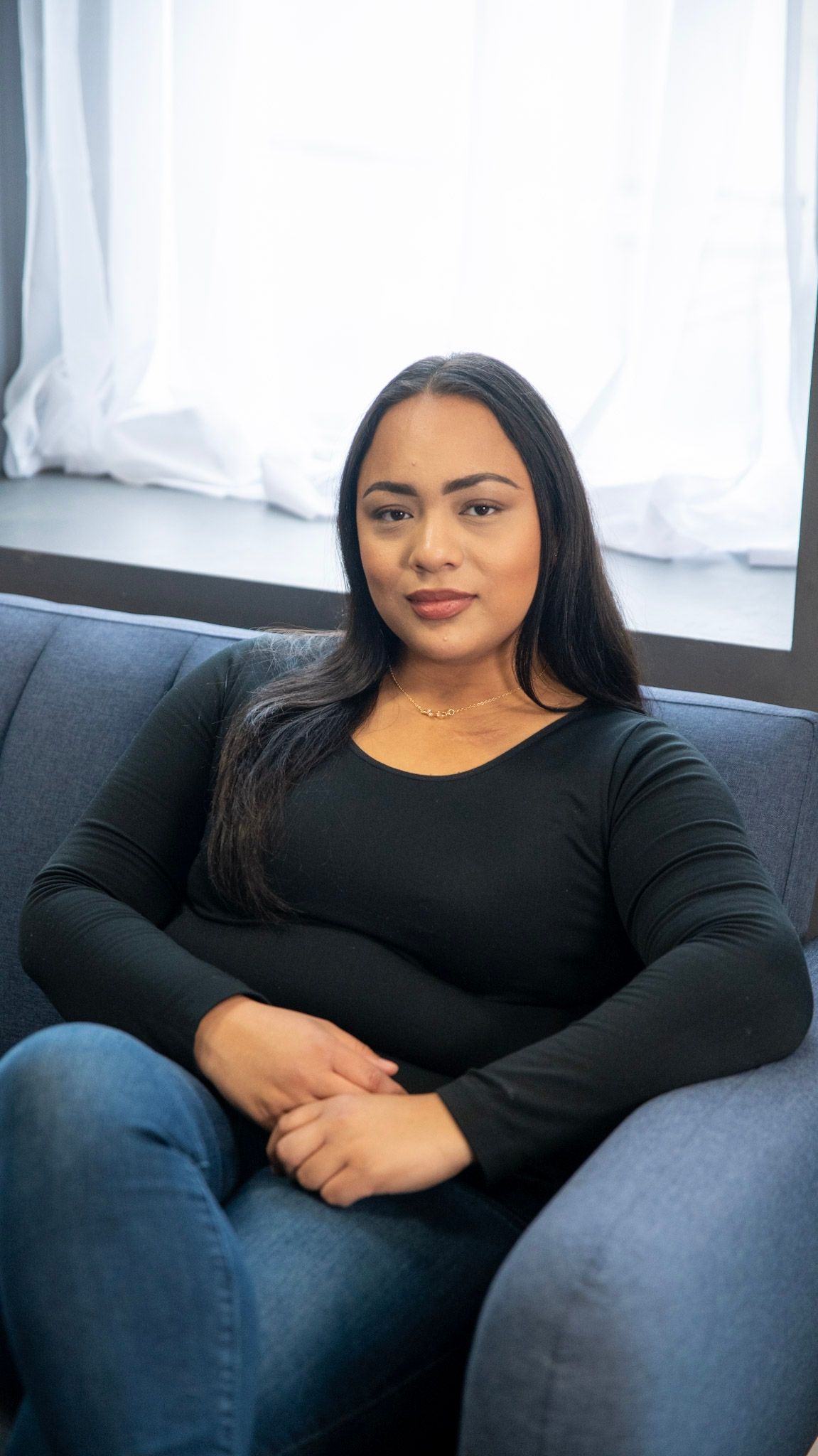 Woman in black long sleeve top relaxing on grey couch in bright room.