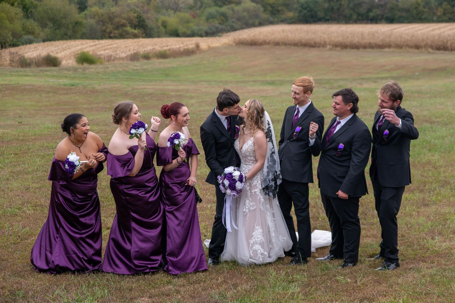 Wedding party lined up in purple and black attire sharing joyful moments in an open field.