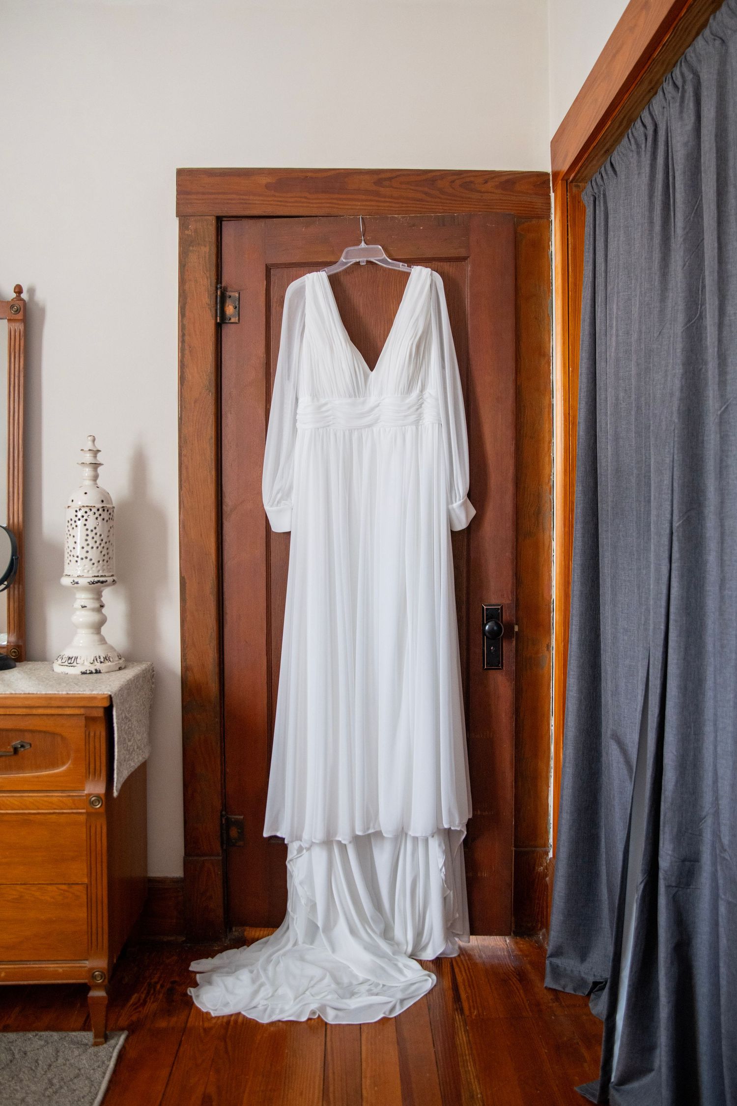 Elegant white wedding dress with long sleeves and flowing train hanging on wooden door in rustic room setting.