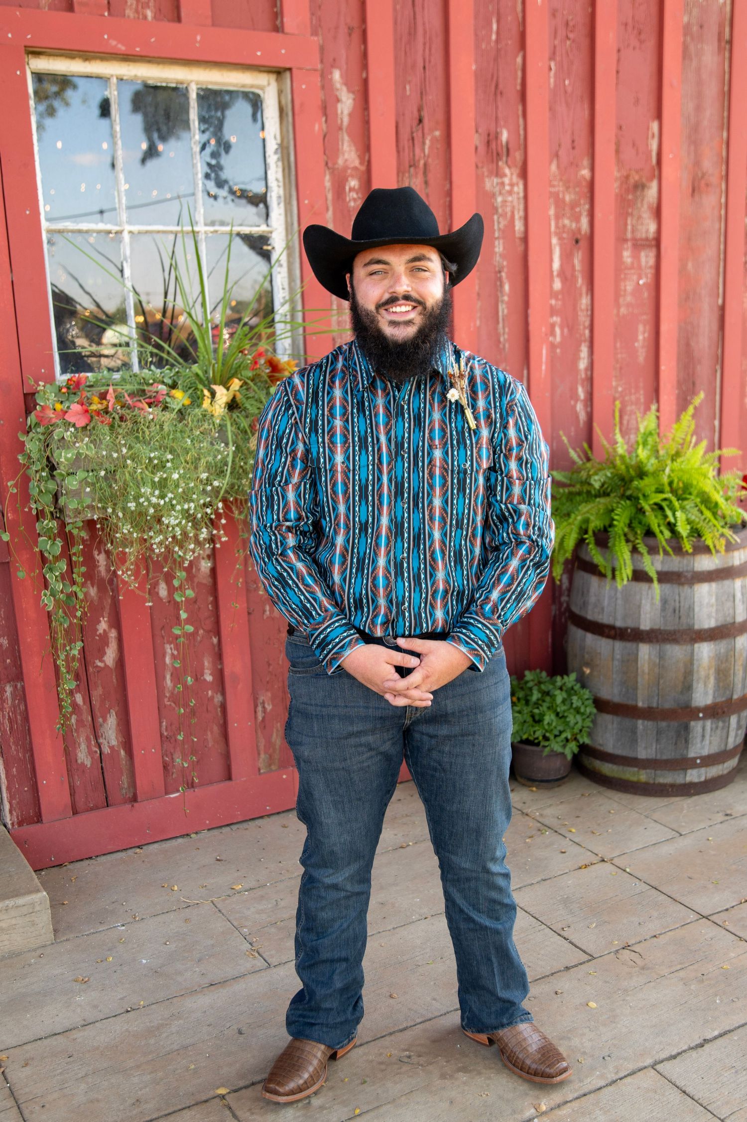 Person in striped blue shirt and cowboy hat poses against red barn wall with barrel planters.