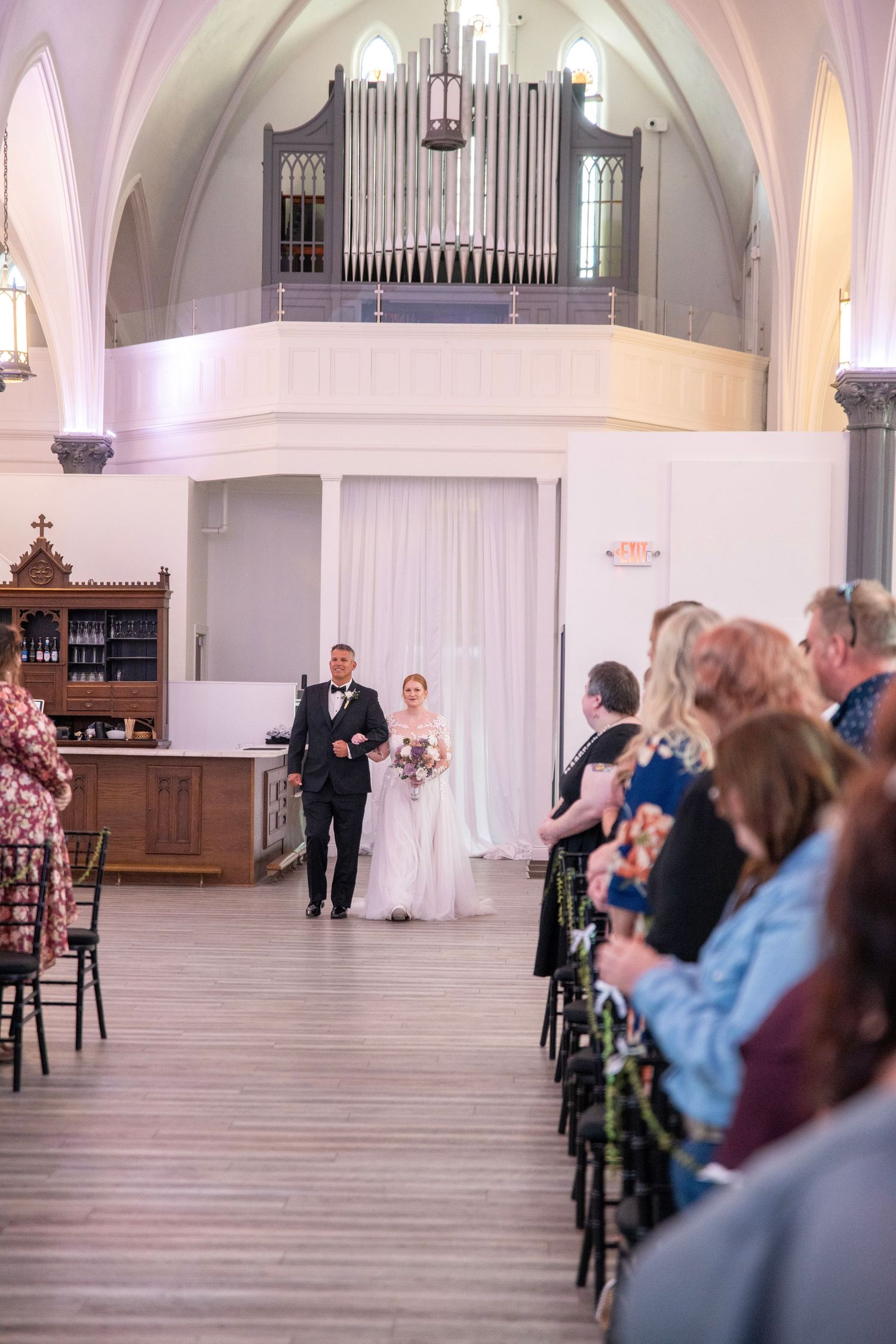 Wedding ceremony in a bright white church with vaulted ceilings and wooden floors.