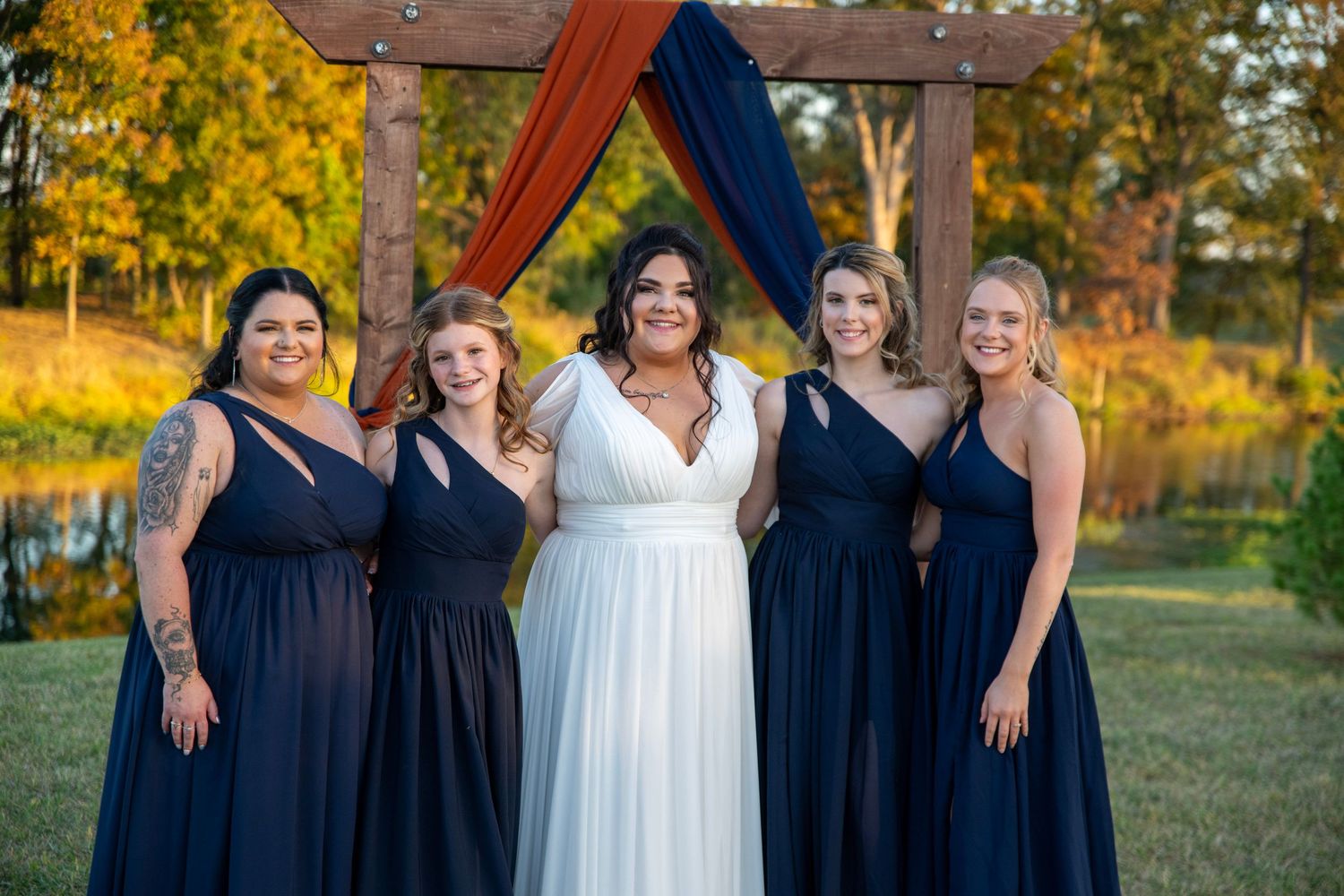Bridesmaids in navy dresses pose with bouquets against rustic wooden arch.