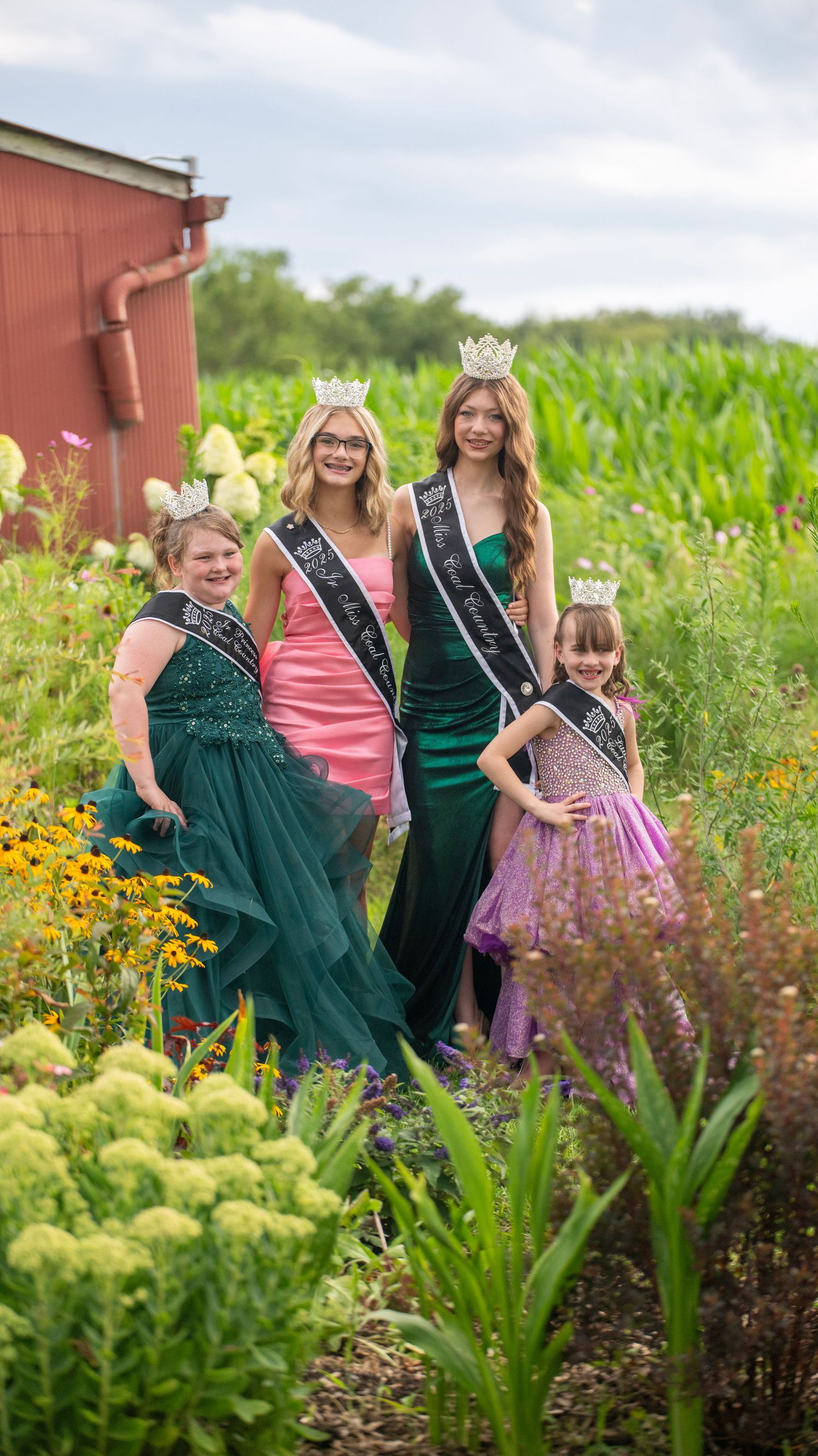 Four people wearing formal attire and crowns pose together in a vibrant garden setting.