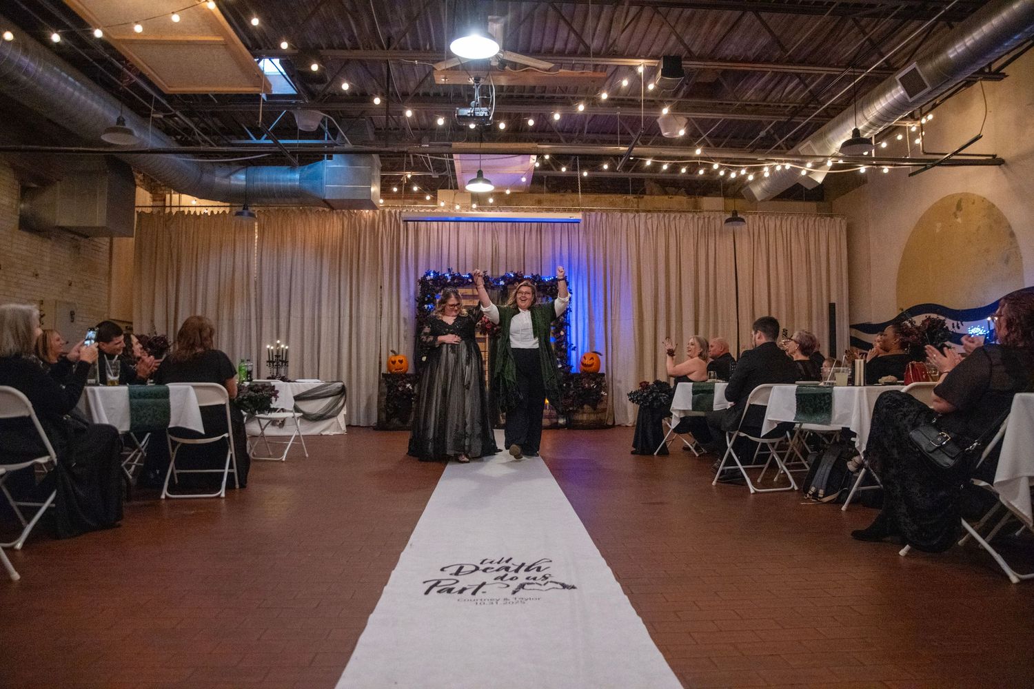 Indoor wedding ceremony setup with white chairs and aisle runner in an industrial-style venue with string lights.
