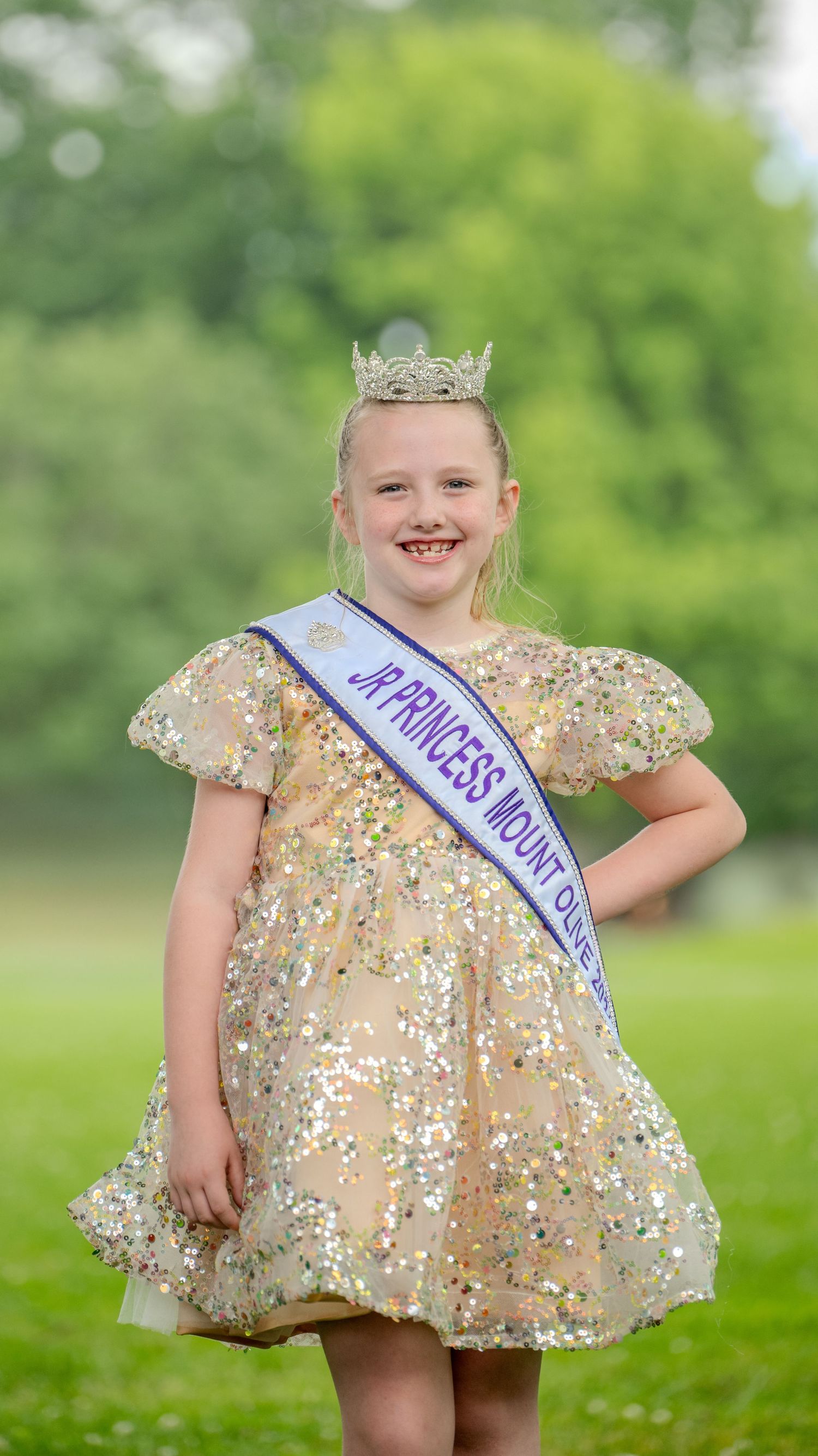 Young pageant contestant in sparkly gold dress with crown and sash smiling outdoors.