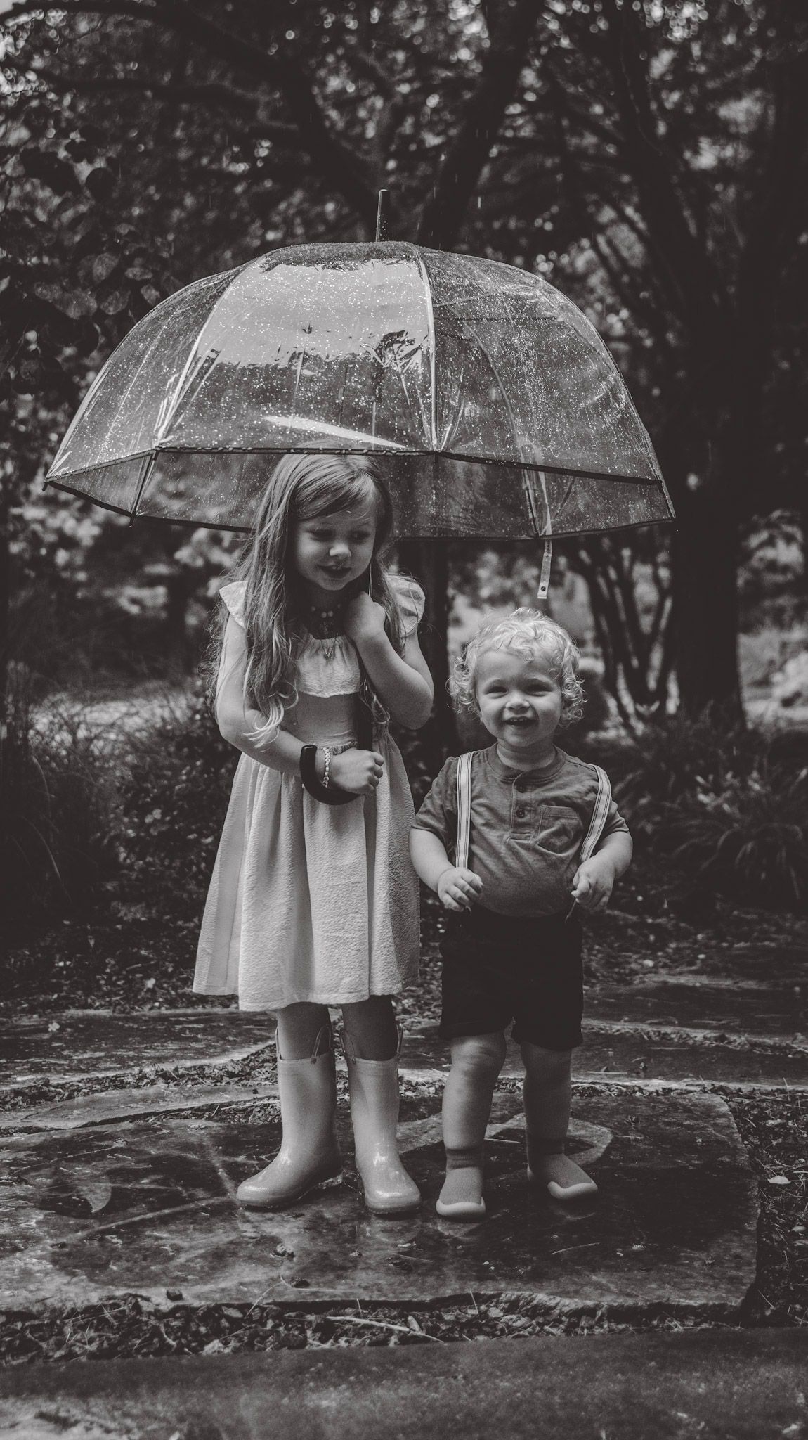 Two children share a clear umbrella while playing in the rain in a garden setting, captured in black and white.