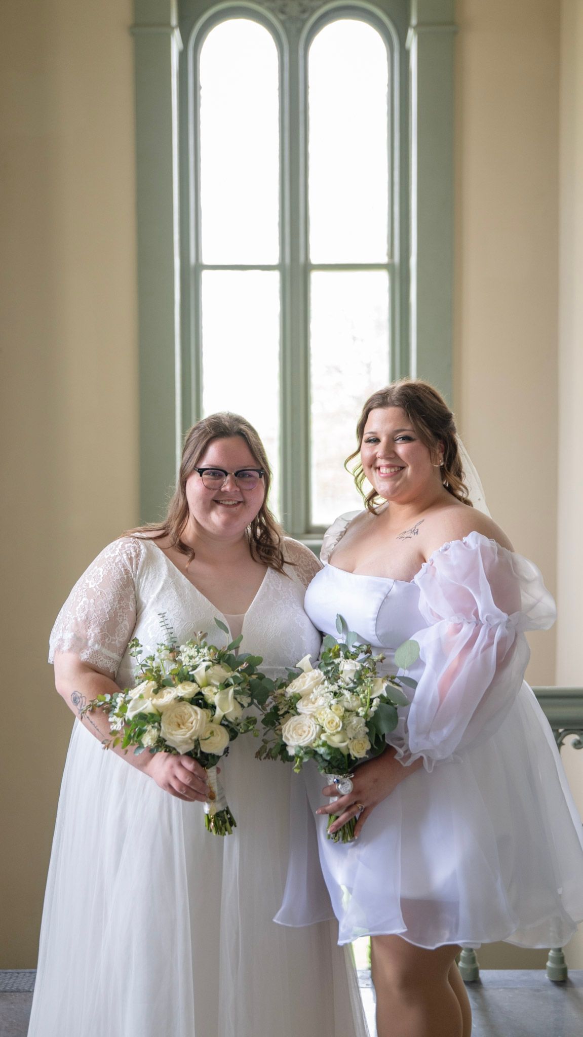 Wedding party in white dresses posing by tall windows in an elegant room with natural light.