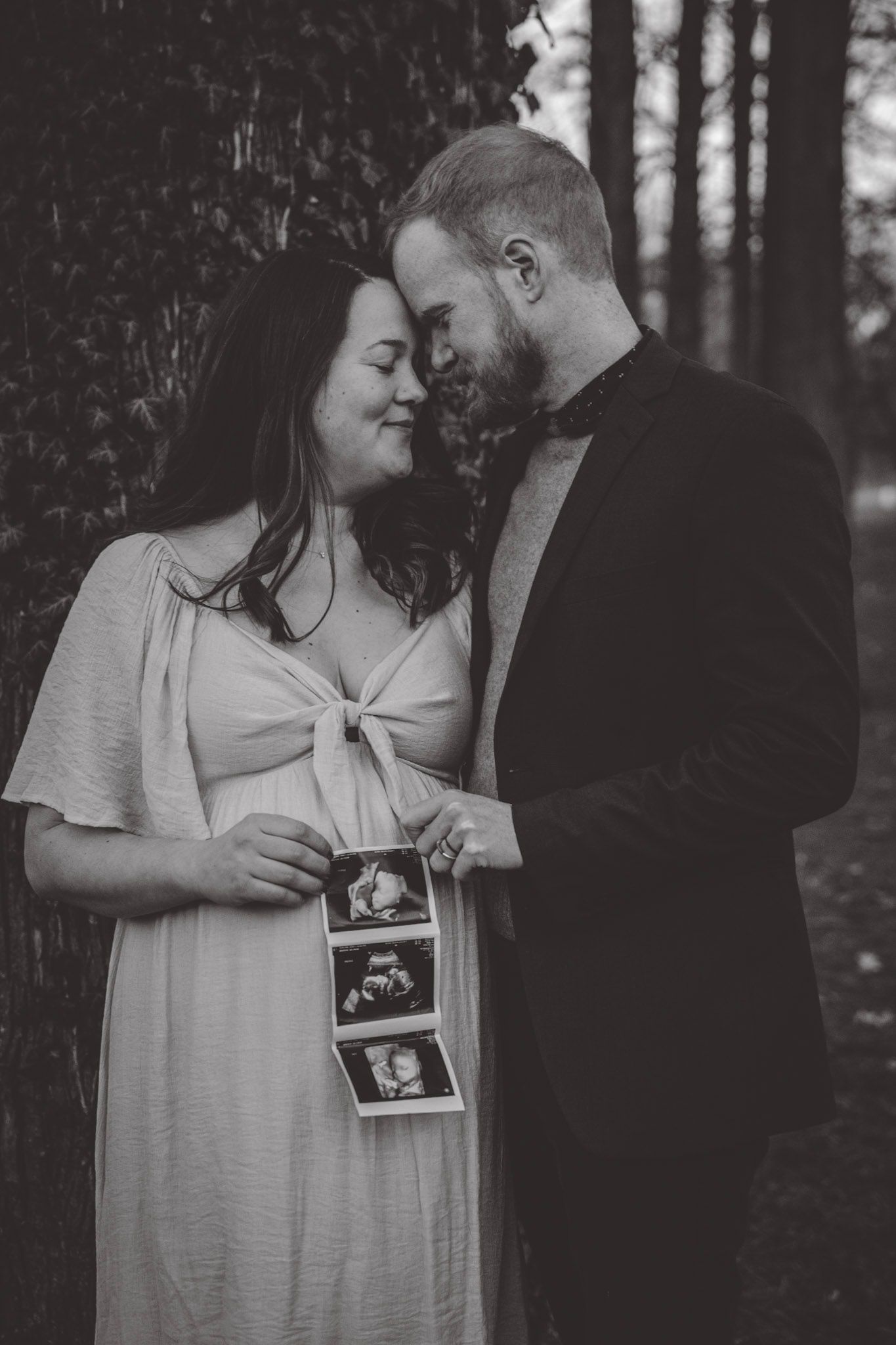 A couple shares an intimate moment while holding ultrasound photos against a tree backdrop.