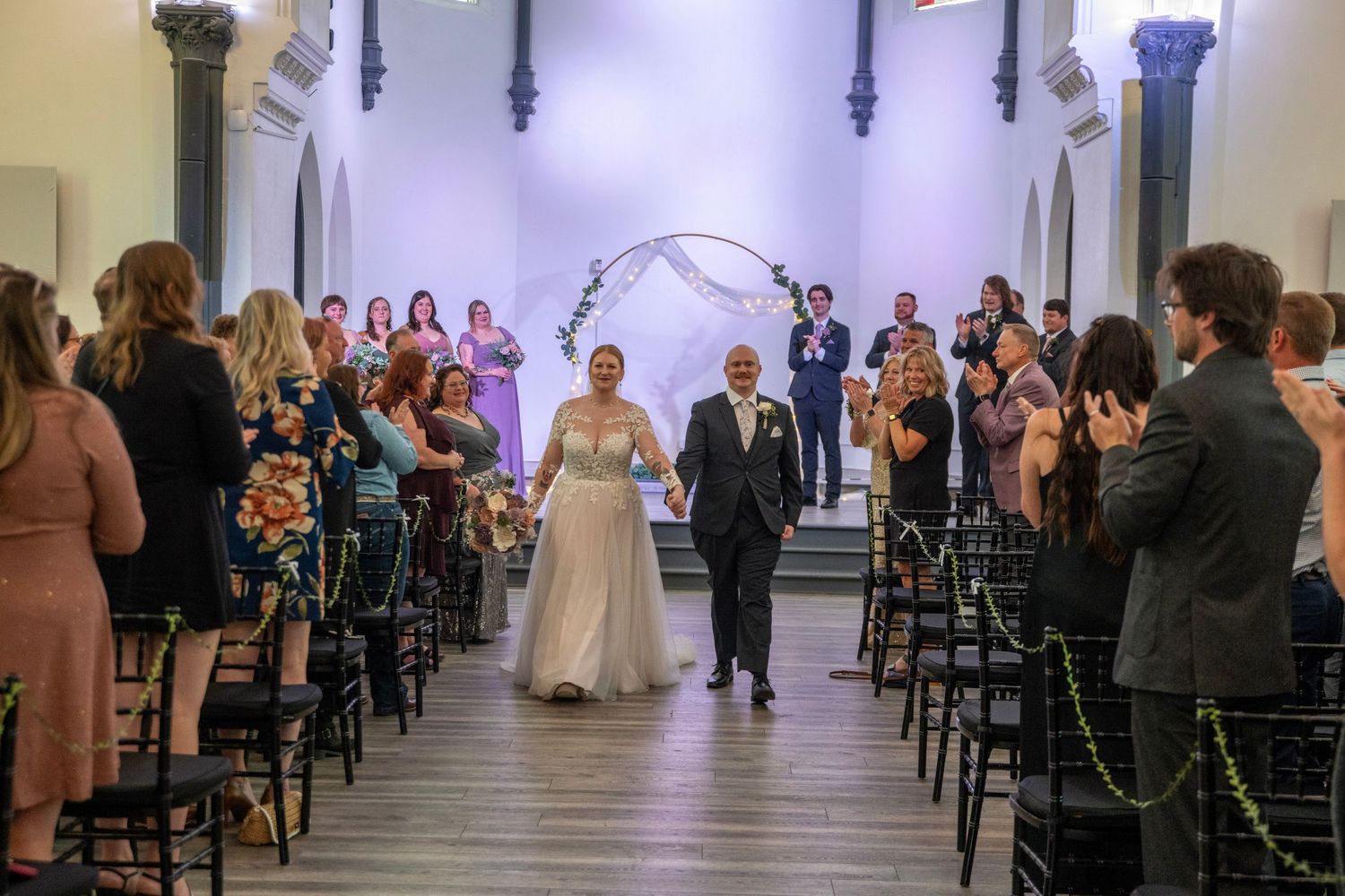 Wedding recessional in chapel with guests standing along aisle as couple walks between wooden chairs.