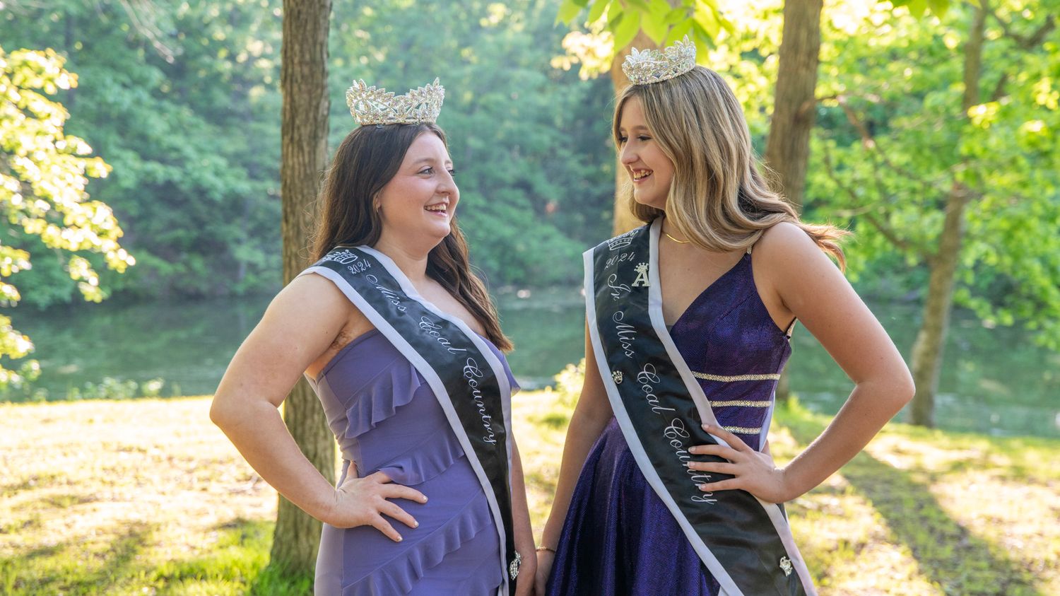 Pageant contestants in evening wear stand together against a backdrop of green trees.