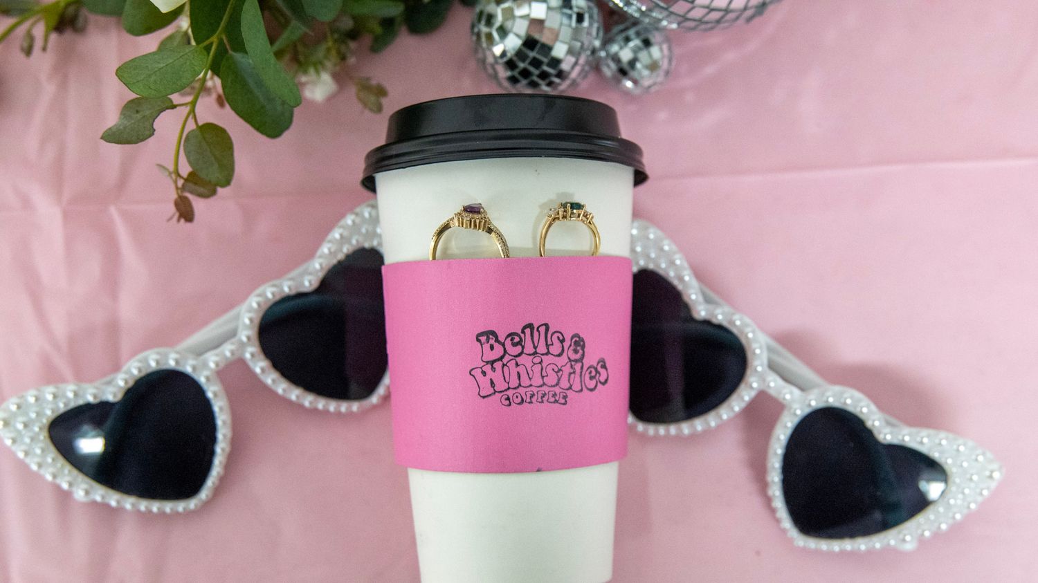 Pink coffee cup sleeve with heart-shaped sunglasses and disco ball decorations on pink background.