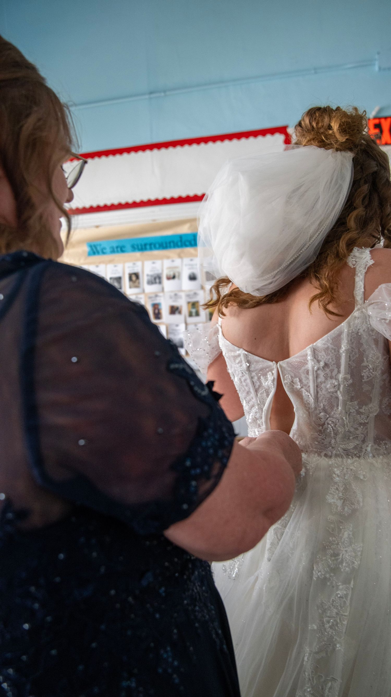 Wedding dress preparation scene with blue wall and decorative border in background.
