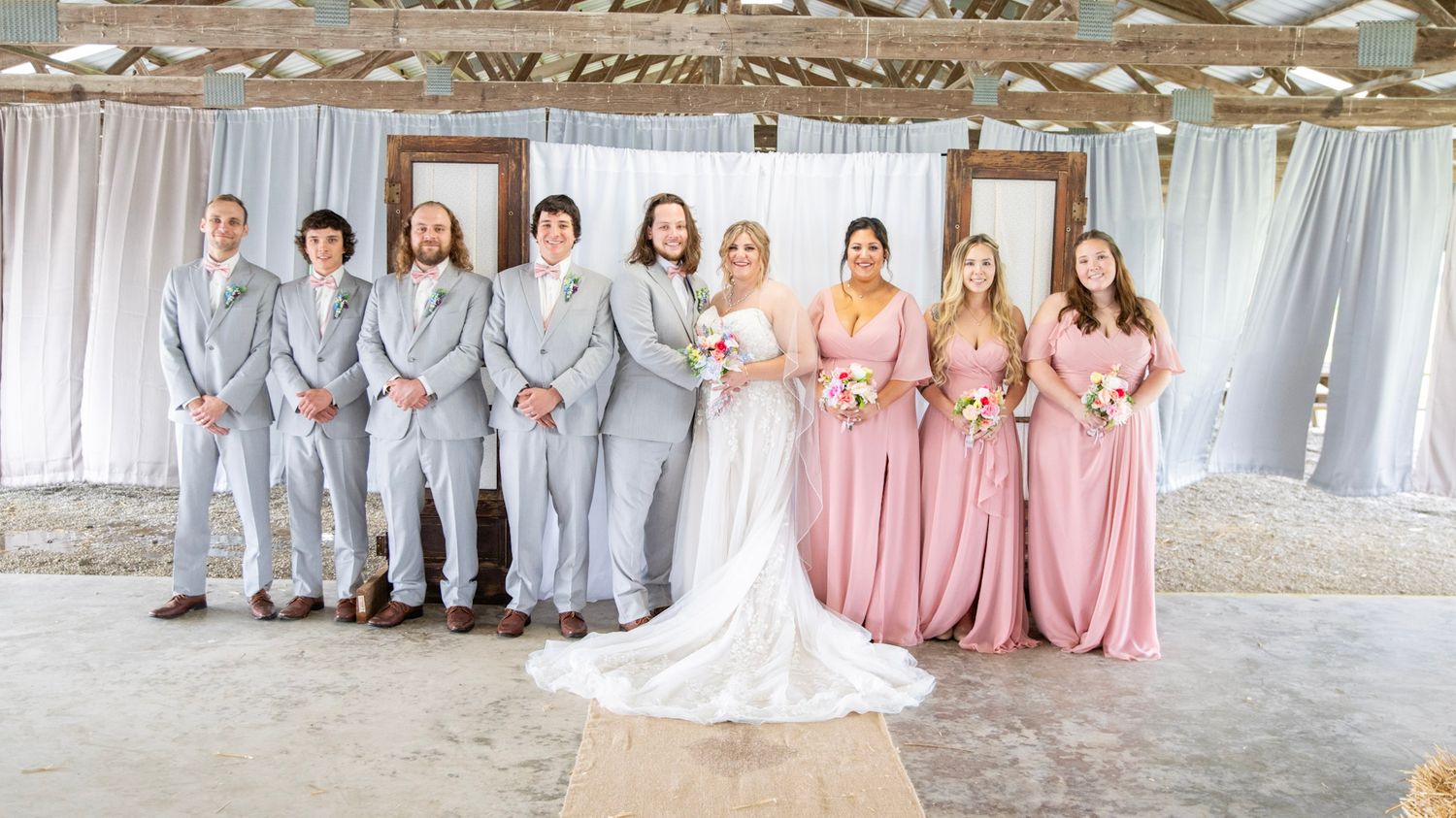 Wedding party members stand in formation wearing coordinated pink dresses and grey suits.