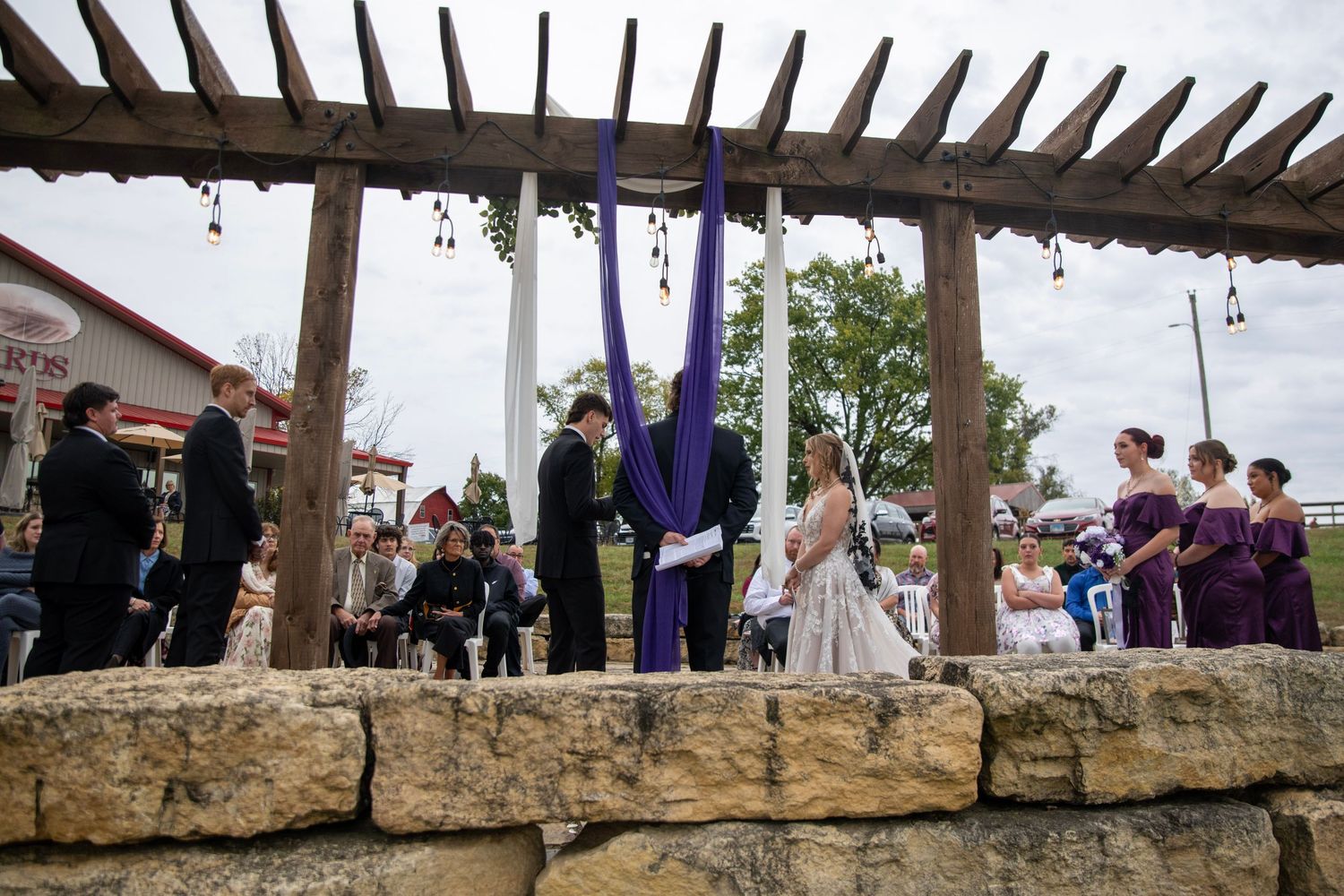 Wedding party gathered under rustic pergola with stone base and hanging decorations.