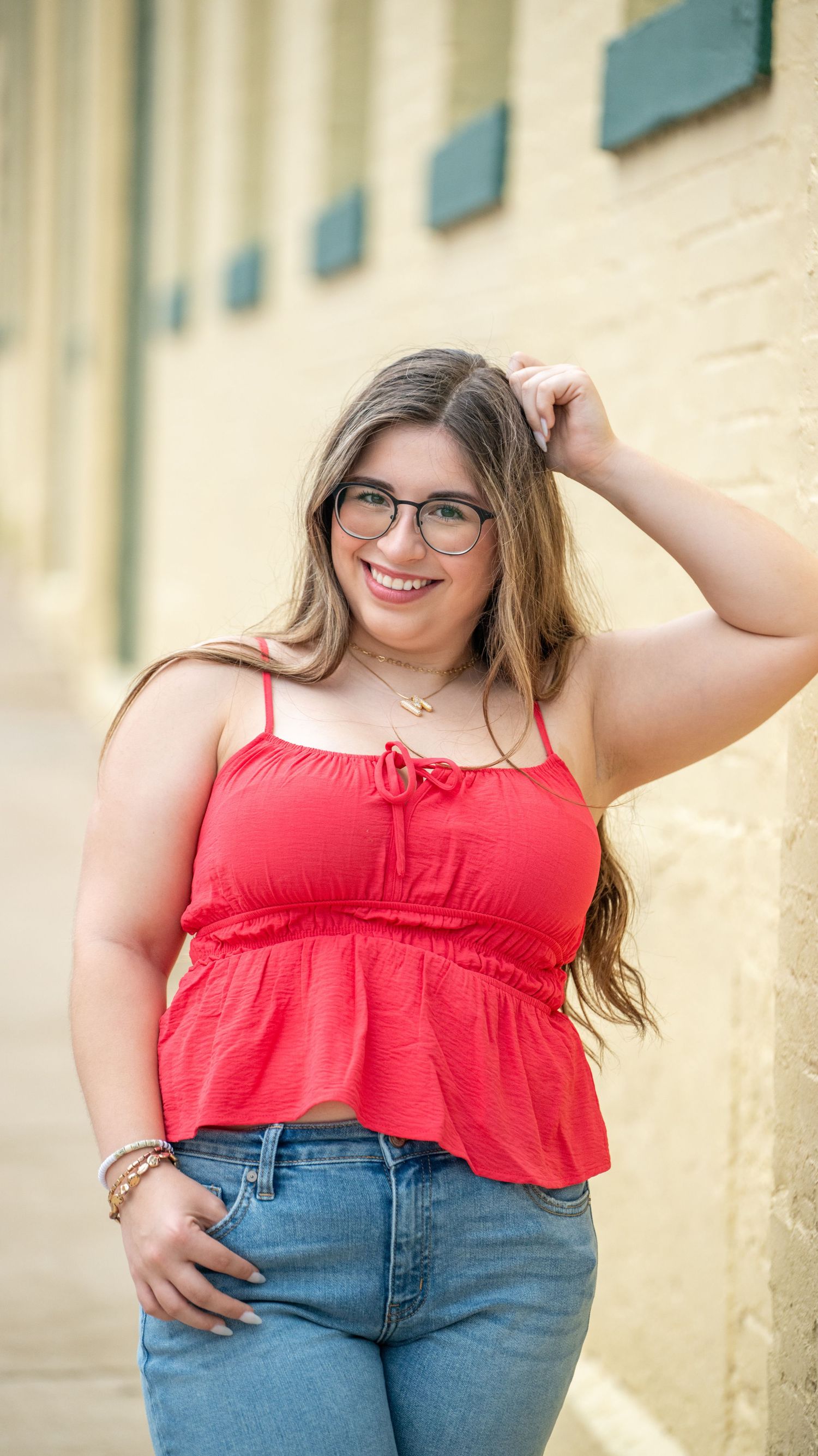 Young woman in red top and jeans poses confidently against cream-colored wall.