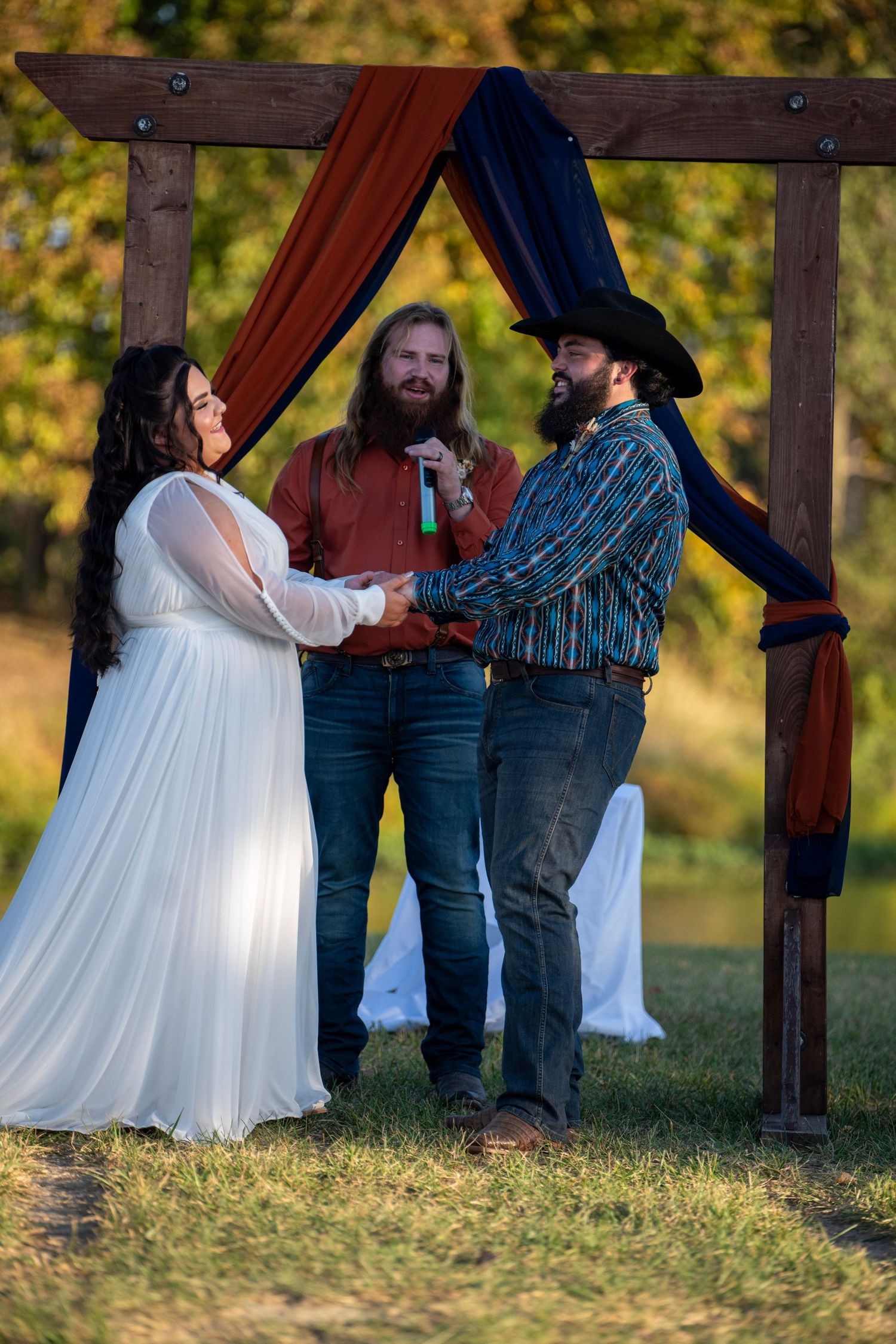 Wedding ceremony under a wooden pergola with autumn foliage in background.