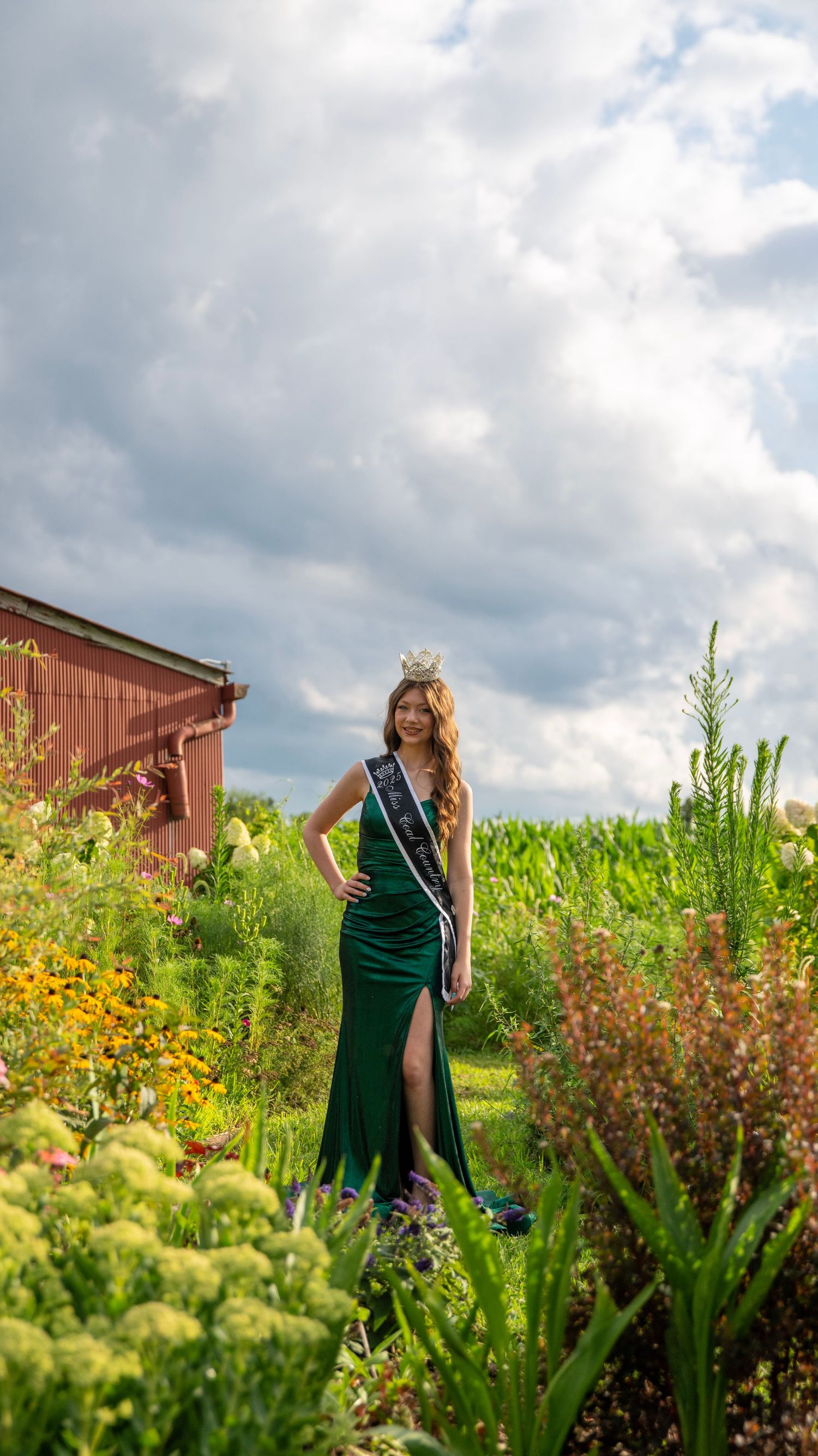 Pageant winner in emerald gown poses in garden setting with red barn backdrop.