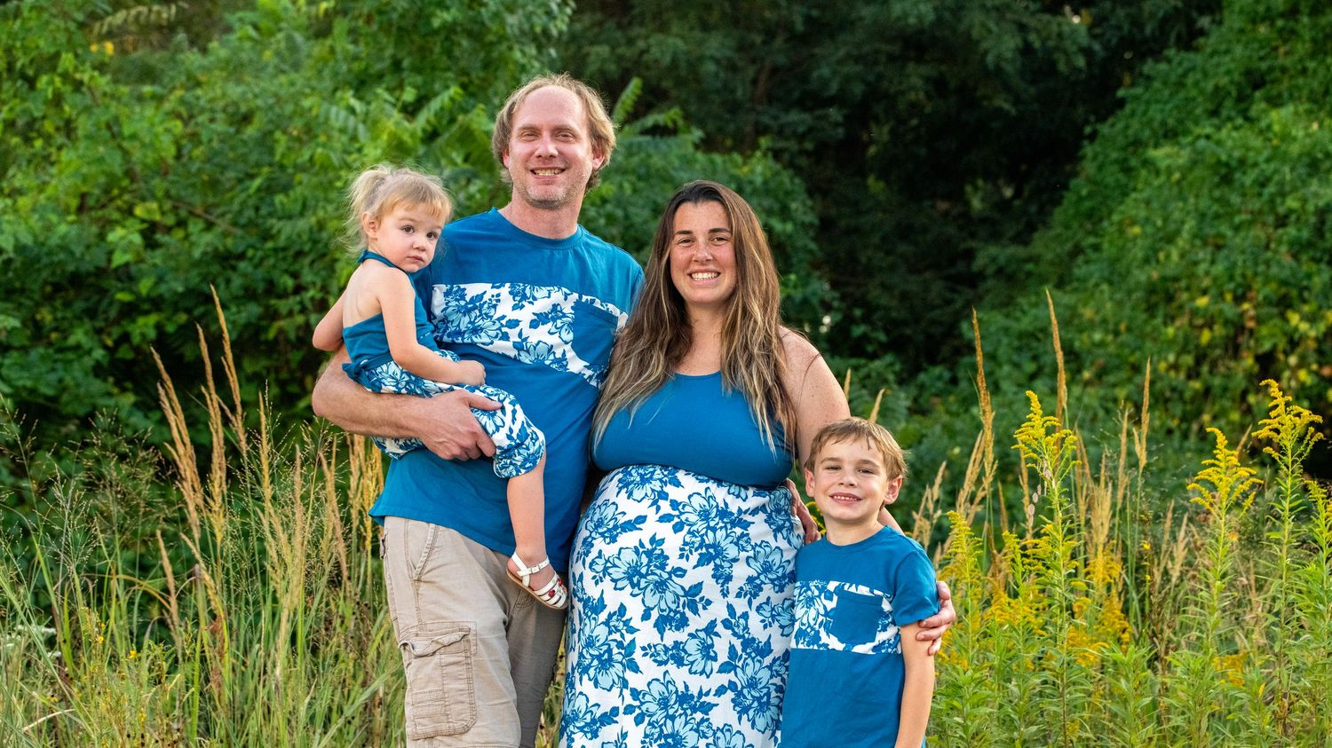 Panoramic family photo session in a lush green field with wildflowers and coordinated blue attire.