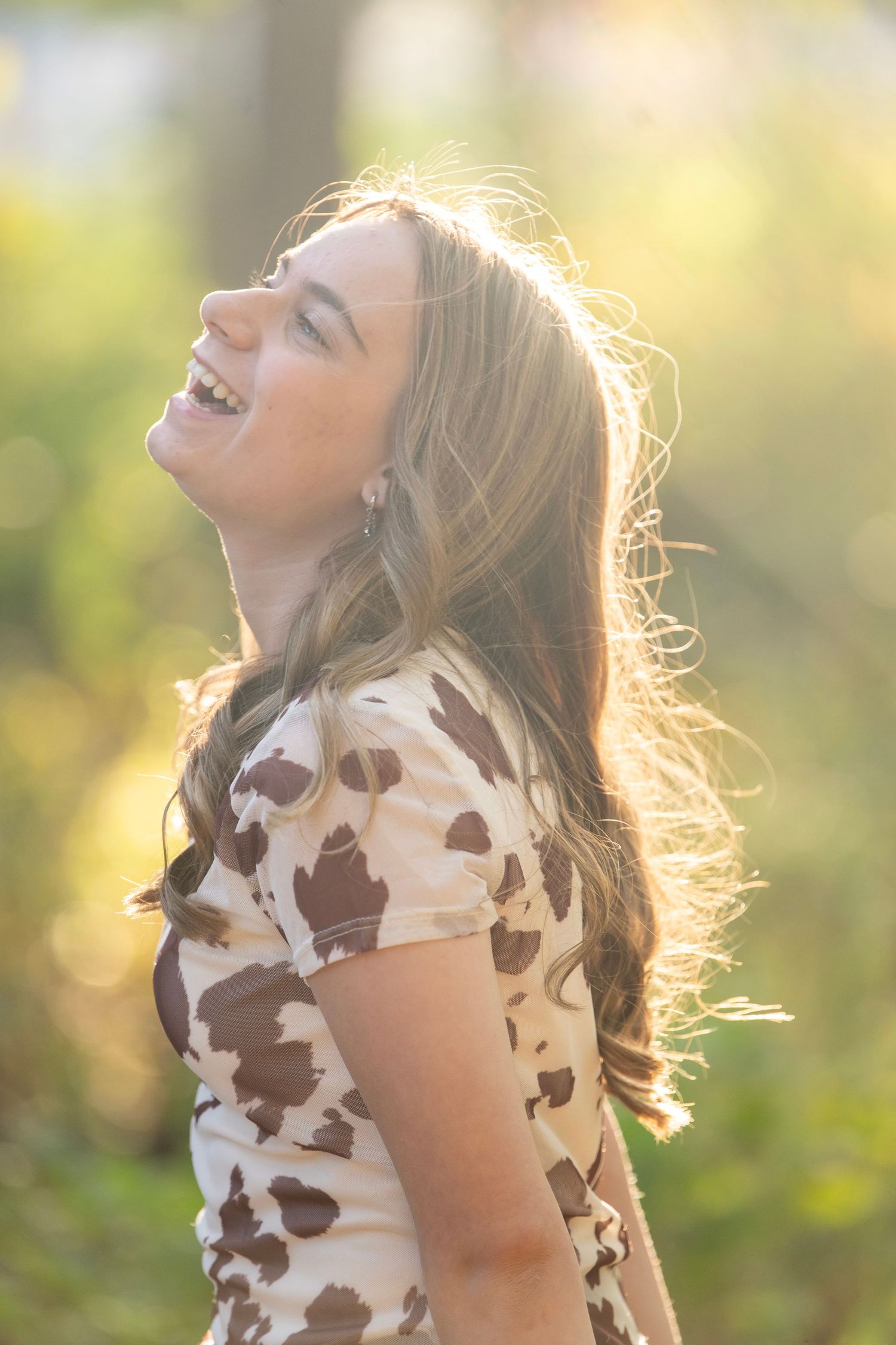 Someone in a brown and white cow print dress poses gracefully in golden sunlight with trees in the background.