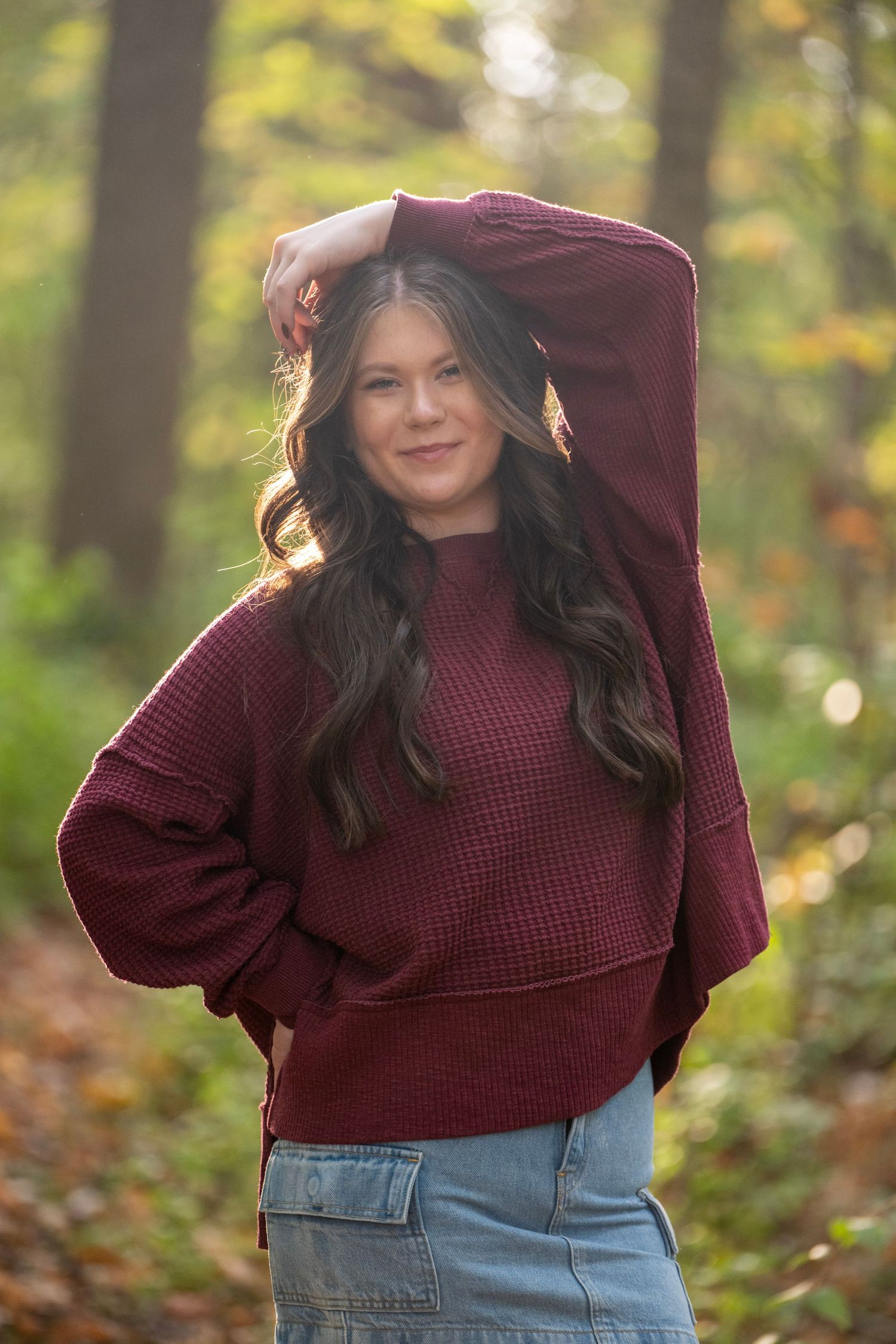 Person in burgundy sweater poses playfully in autumn forest setting.