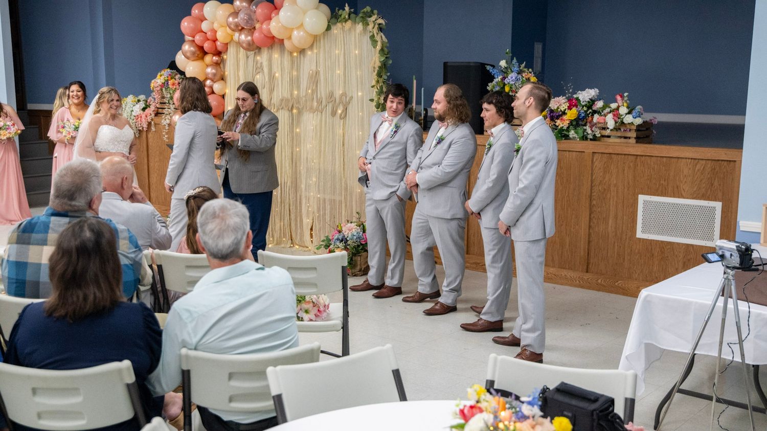 Wedding ceremony in progress with light gray suited wedding party and guests in white chairs.
