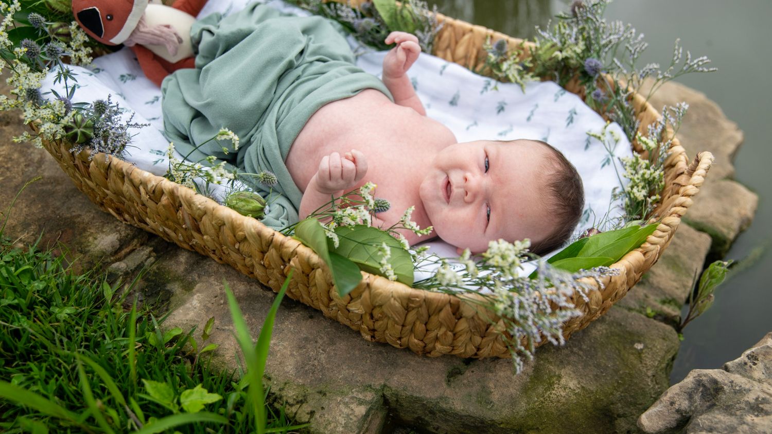 A happy baby lies in a woven basket decorated with fresh flowers and greenery.