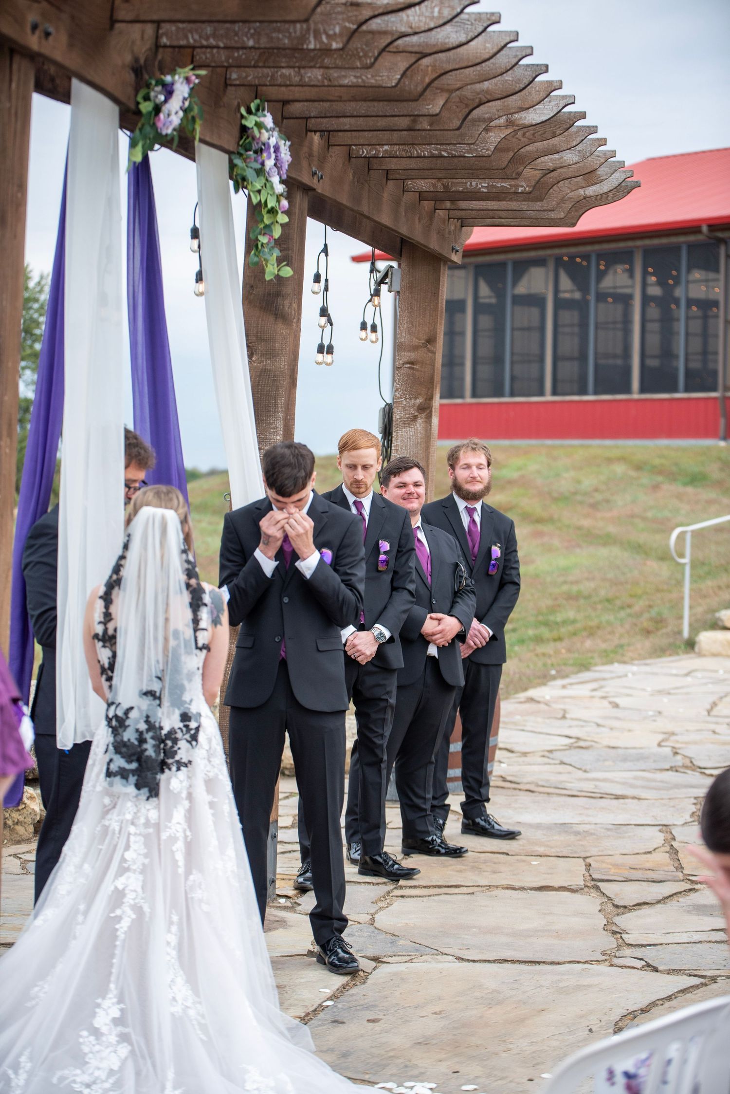Wedding ceremony under wooden pergola decorated with purple and white drapes and floral accents.