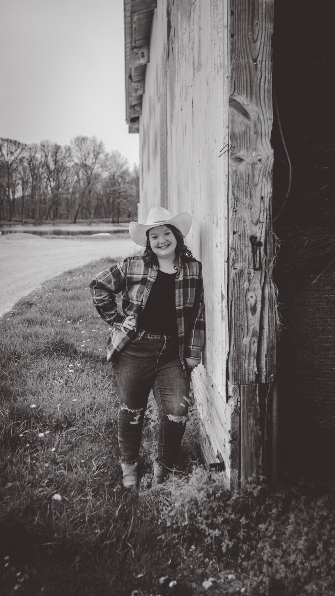 Person in plaid shirt poses against rustic wooden wall in black and white photo.