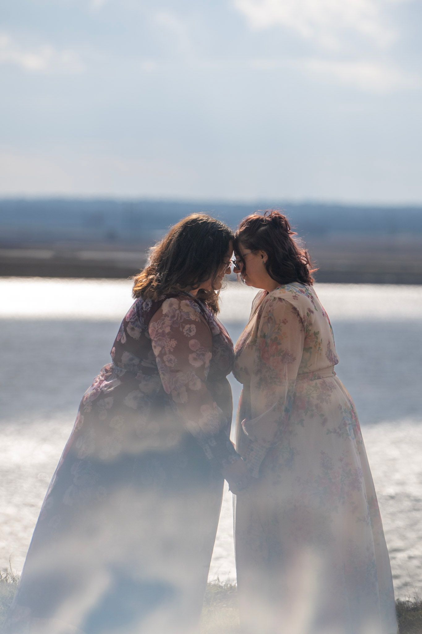 Dreamy beach photoshoot with two people in flowy dresses against sunlit water.