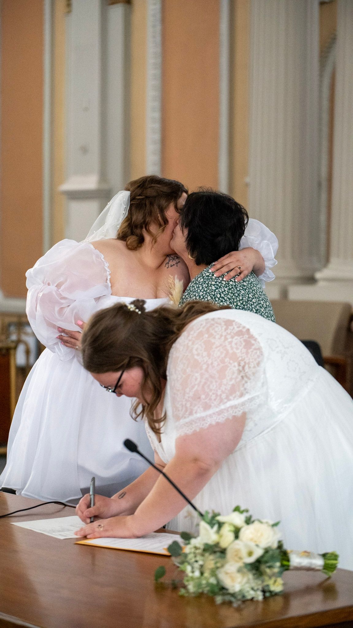 Two people in wedding dresses signing marriage documents at courthouse.