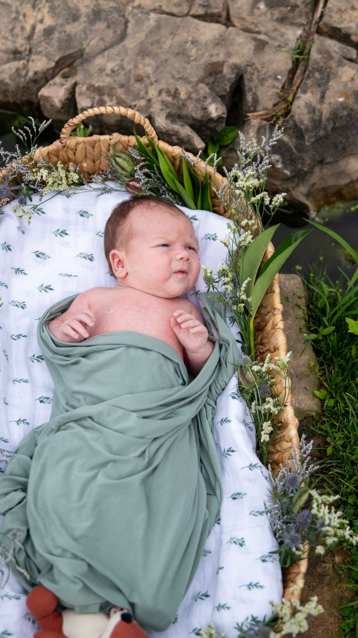 A content baby rests in a decorated basket with sage green wrapping and floral accents.